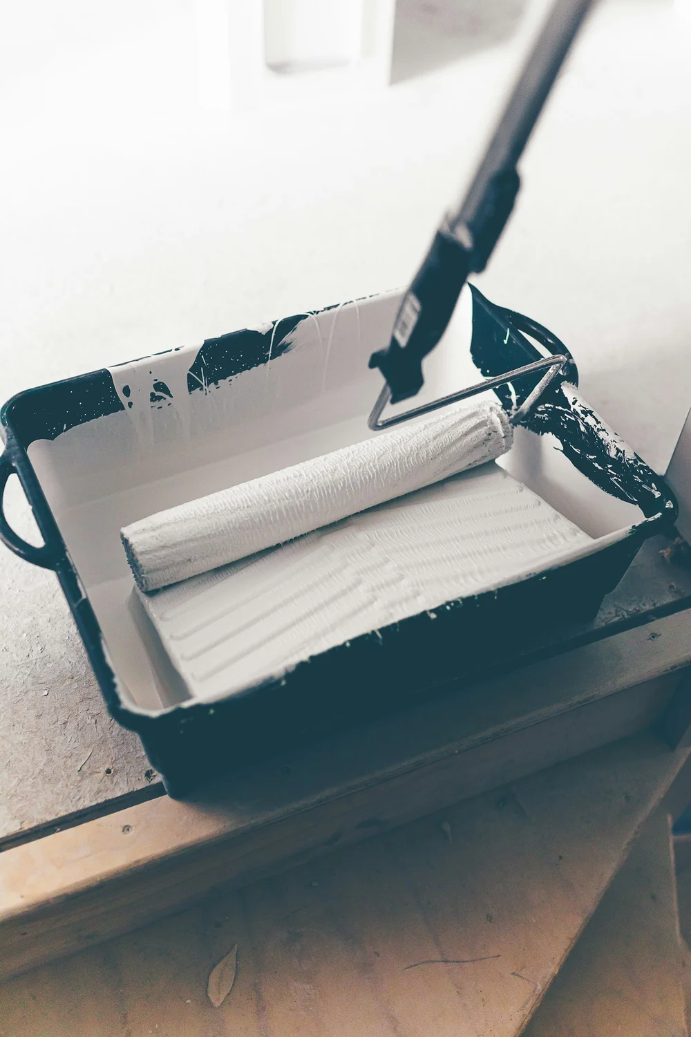 A roller on an extension pole loaded with white paint, resting in a tray on a wooden floor