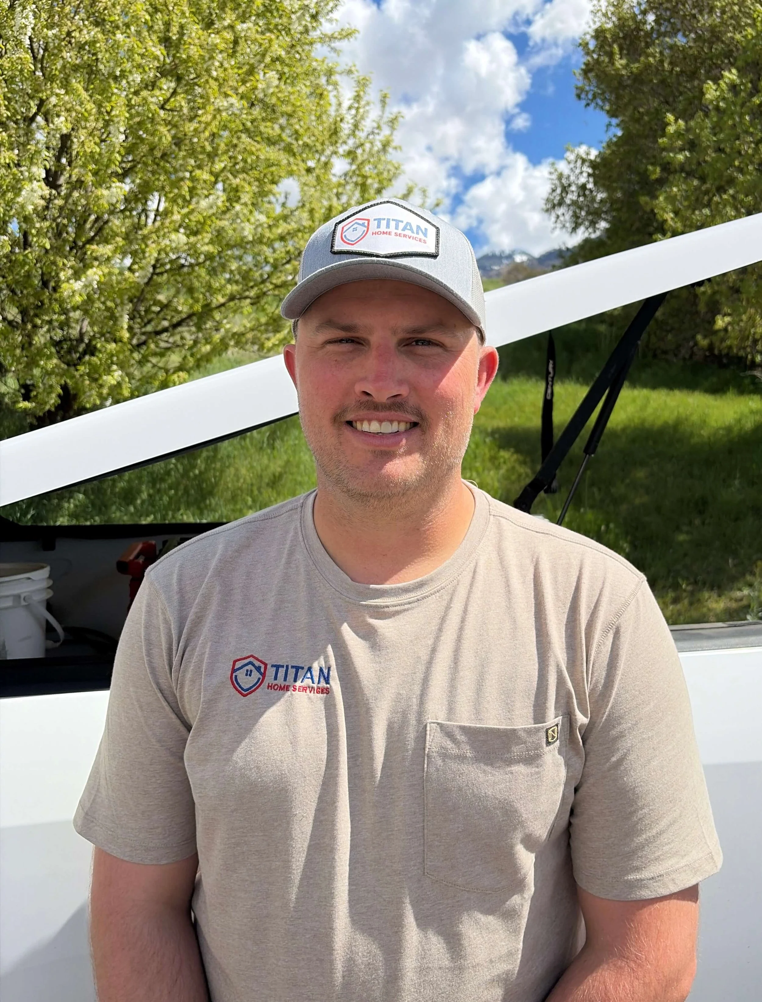 Brendan Michael wearing a gray cap with a Titan Home Services logo and a beige T-shirt, standing outdoors in front of a white vehicle and greenery, under a partly cloudy sky.