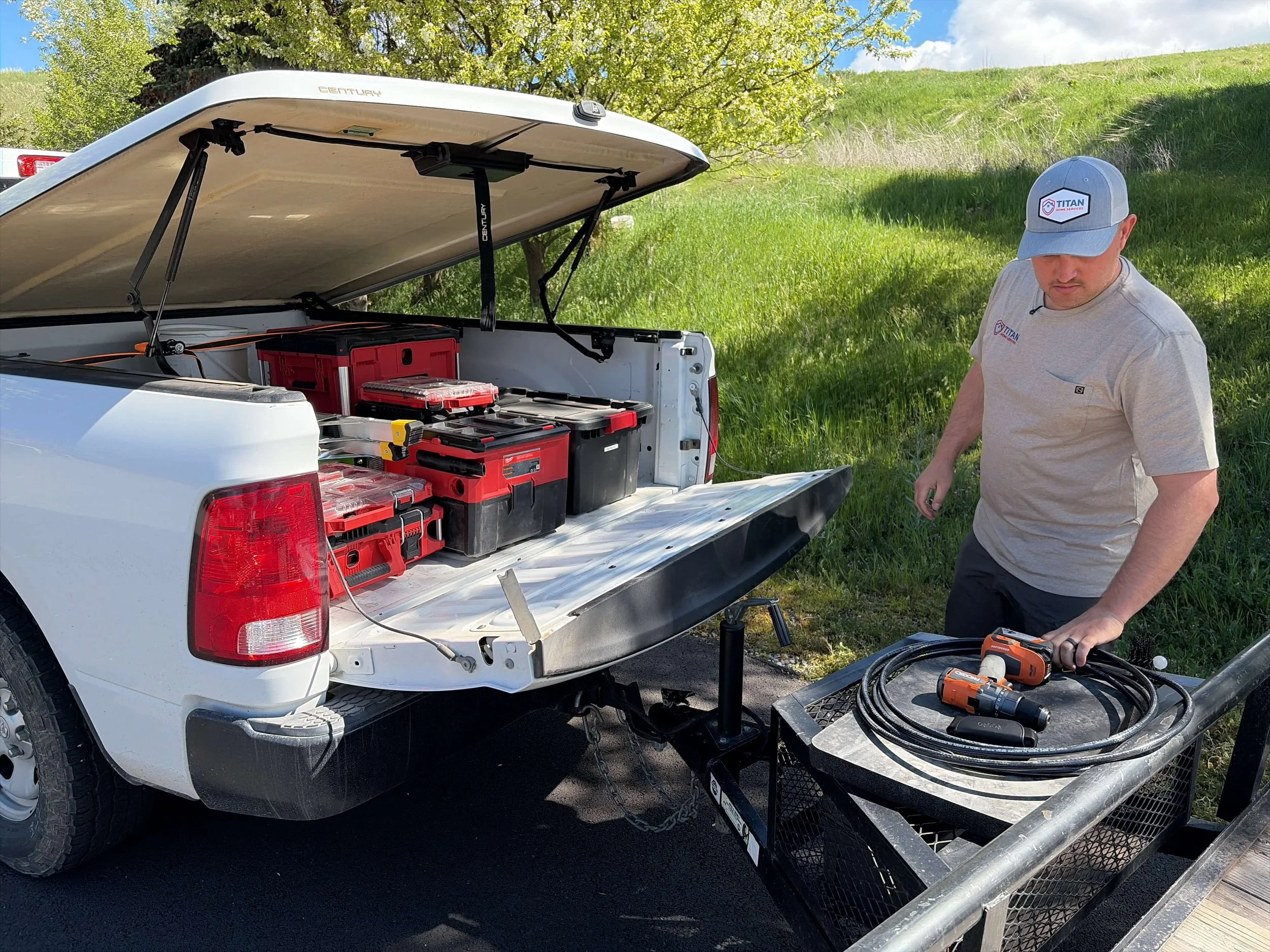 Handyman working with power tools near open pickup truck with toolbox and equipment on a sunny grassy roadside.