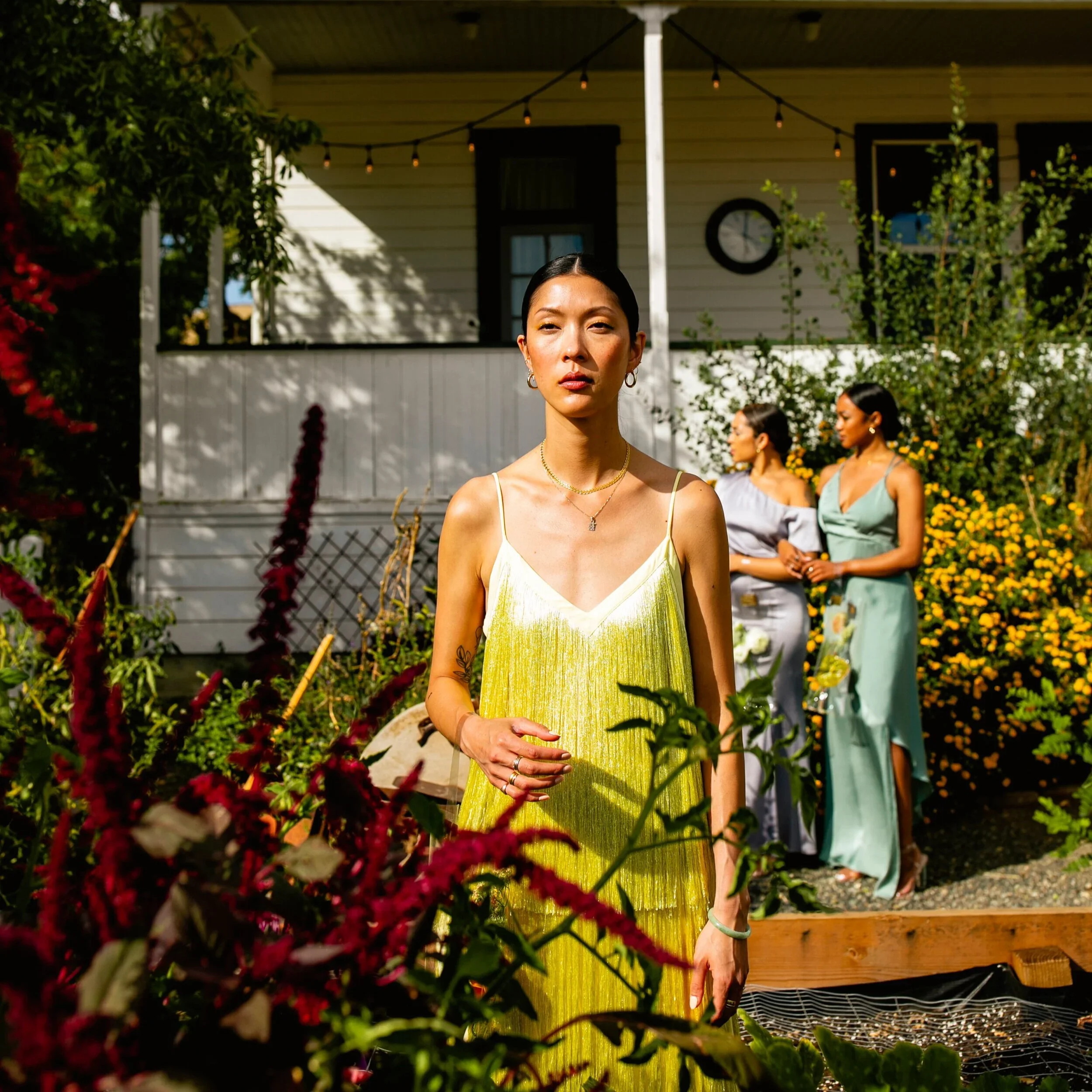 A woman with dark hair and gold jewelry standing outdoors during daytime, with several other women in the background, near a yellow house with hanging string lights and greenery.