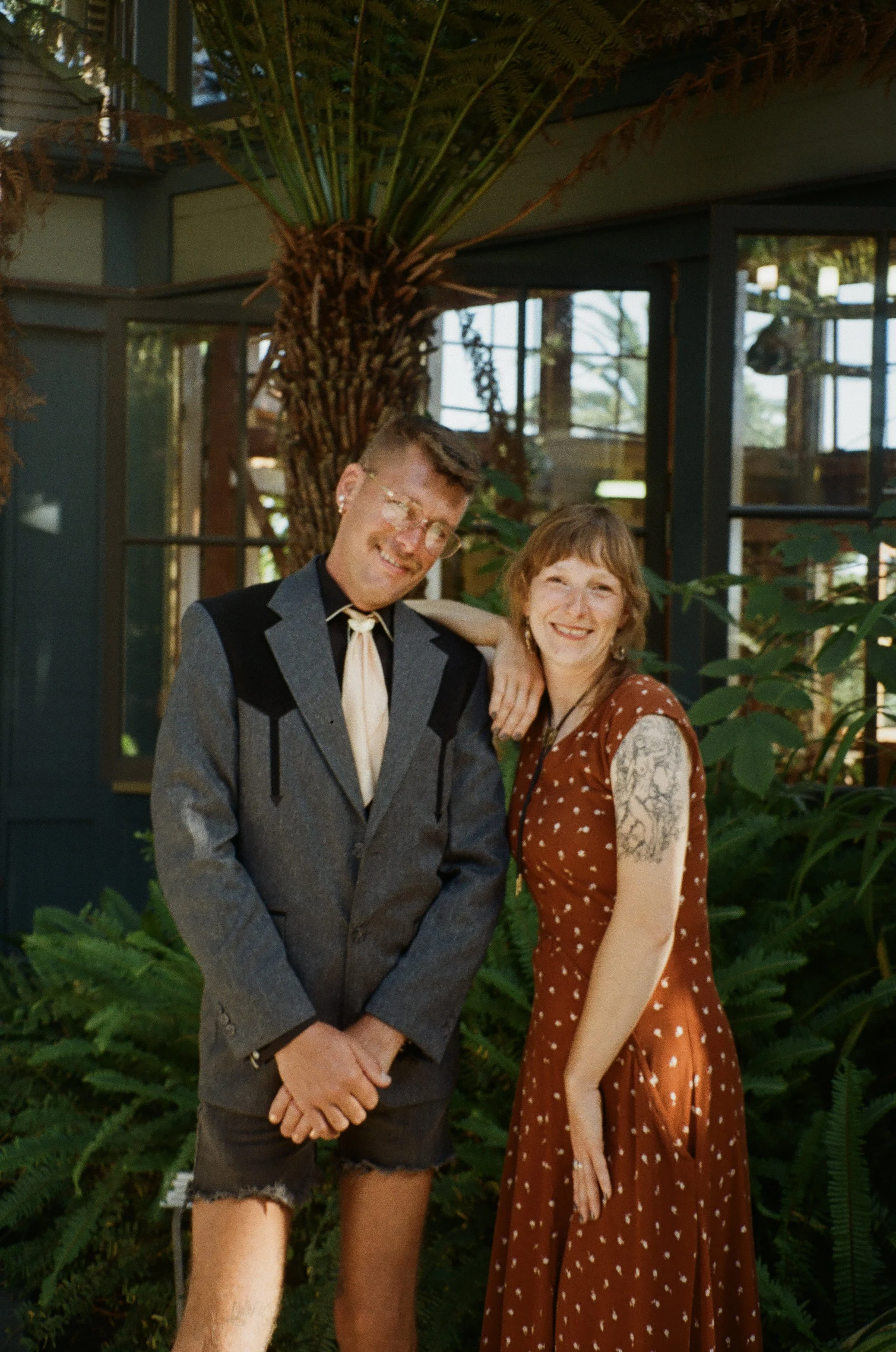 A young man and woman standing together outdoors, smiling, with a large fern plant behind them and modern glass and wood building in the background.