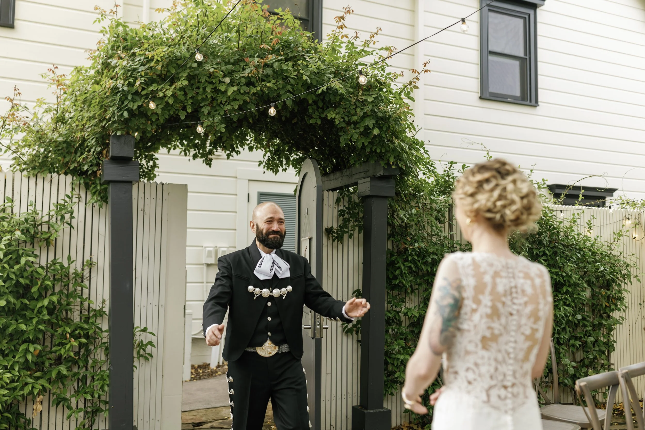 A man dressed as a traditional mariachi with a black suit, silver buttons, and white embroidery, dancing with a woman in a lace dress, outdoors in front of a white fence and greenery, under string lights.