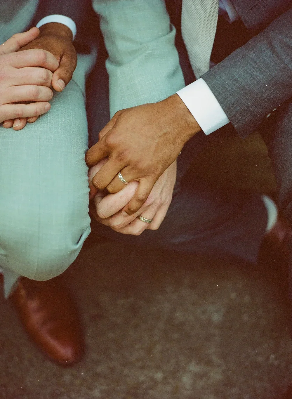 Close-up of hands, one with a wedding ring, gently holding another person's hand, who is dressed in a light-colored suit.