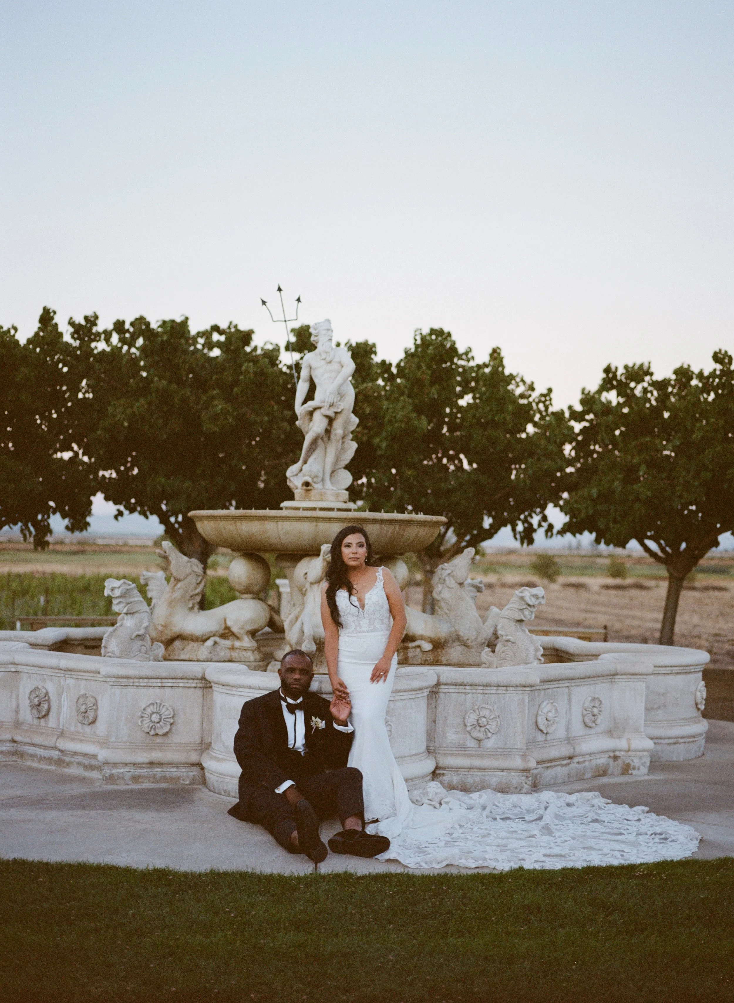 A couple in wedding attire poses in front of a large ornate fountain with a statue of a man and a woman on top, surrounded by trees and open land, during sunset or dusk.