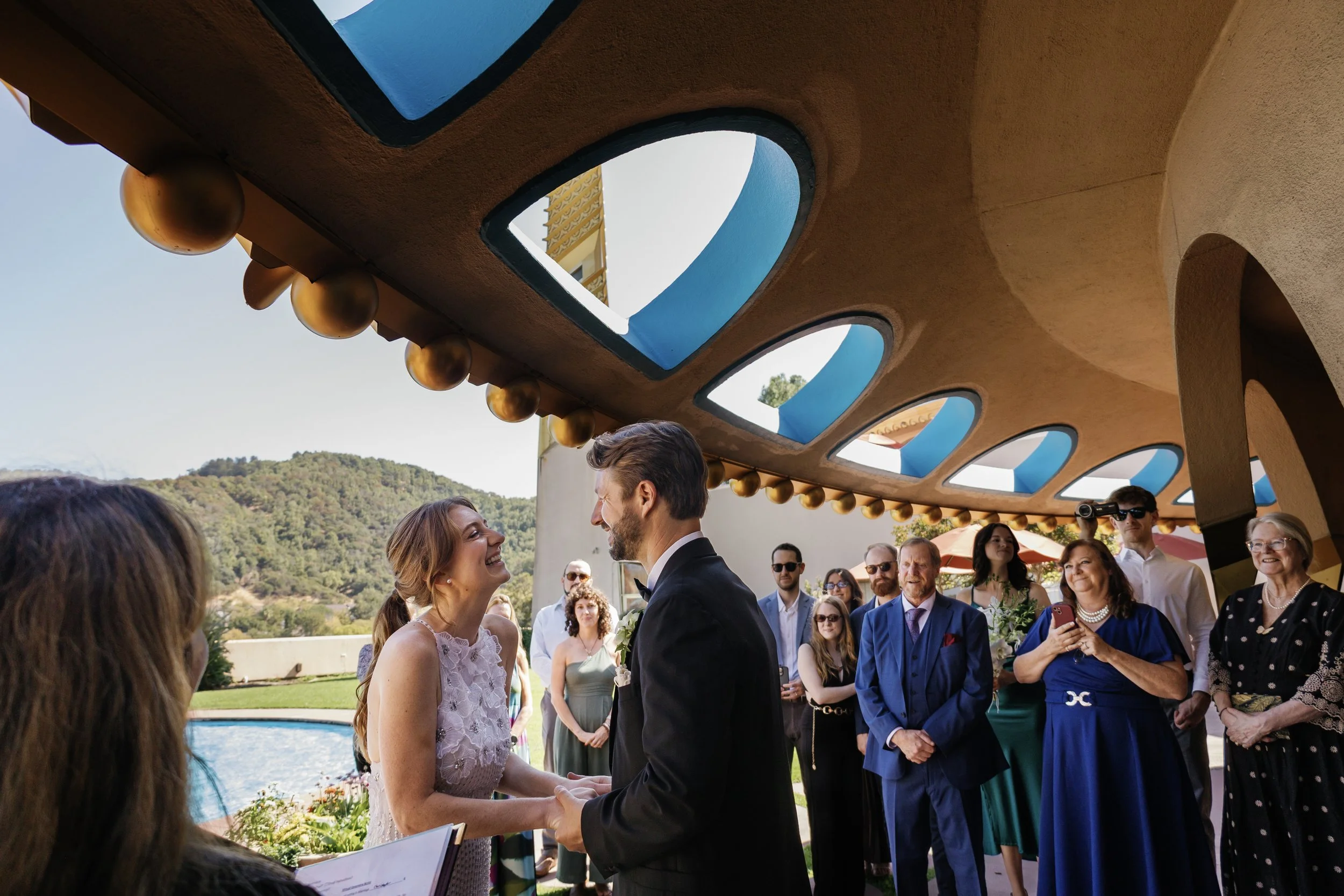 A wedding ceremony with a couple exchanging vows outdoors, surrounded by guests and a decorative ceiling with blue and gold accents.