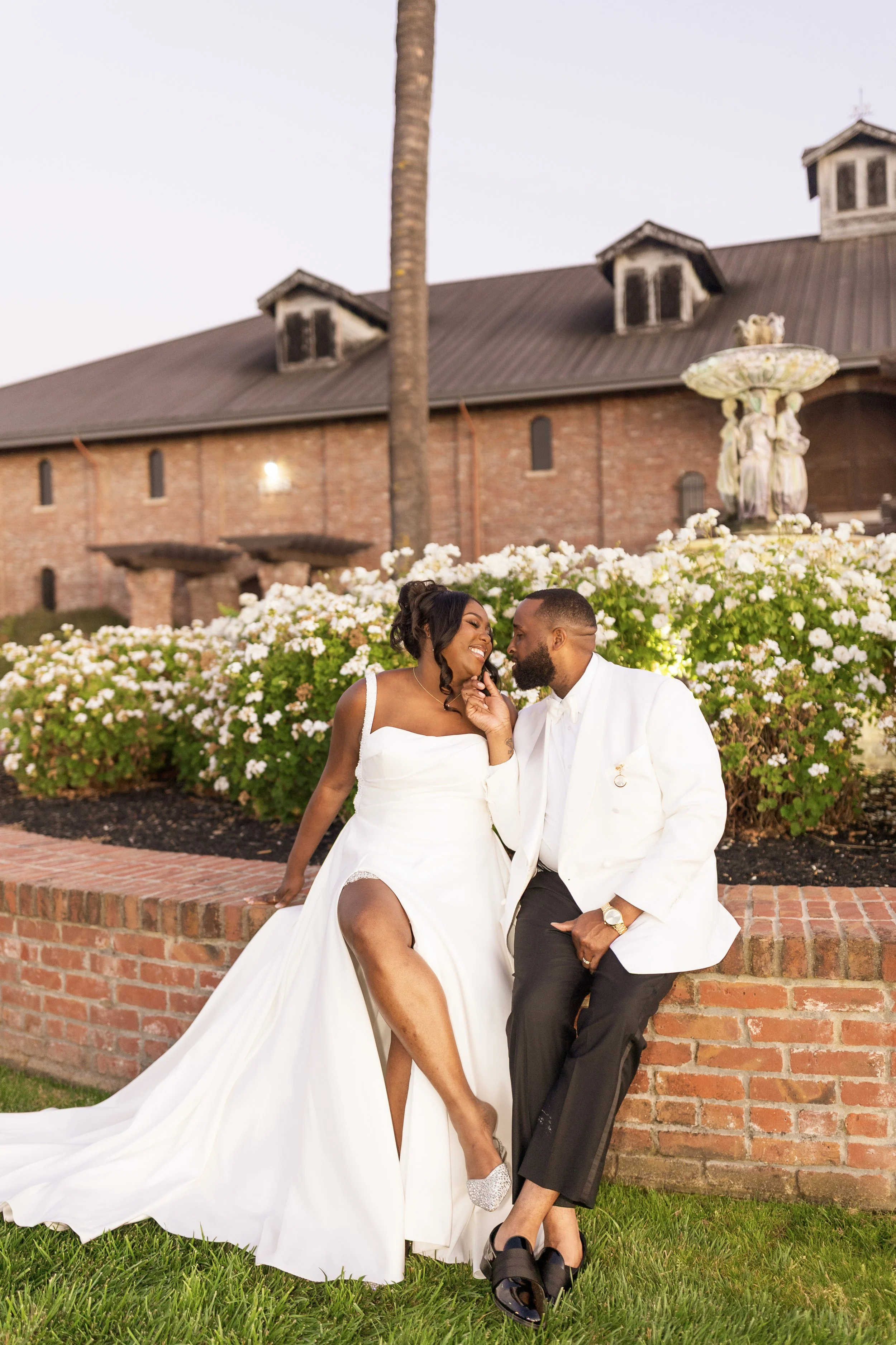A couple dressed in wedding attire sitting on a brick ledge surrounded by white flowers, with a brick building and a fountain in the background.
