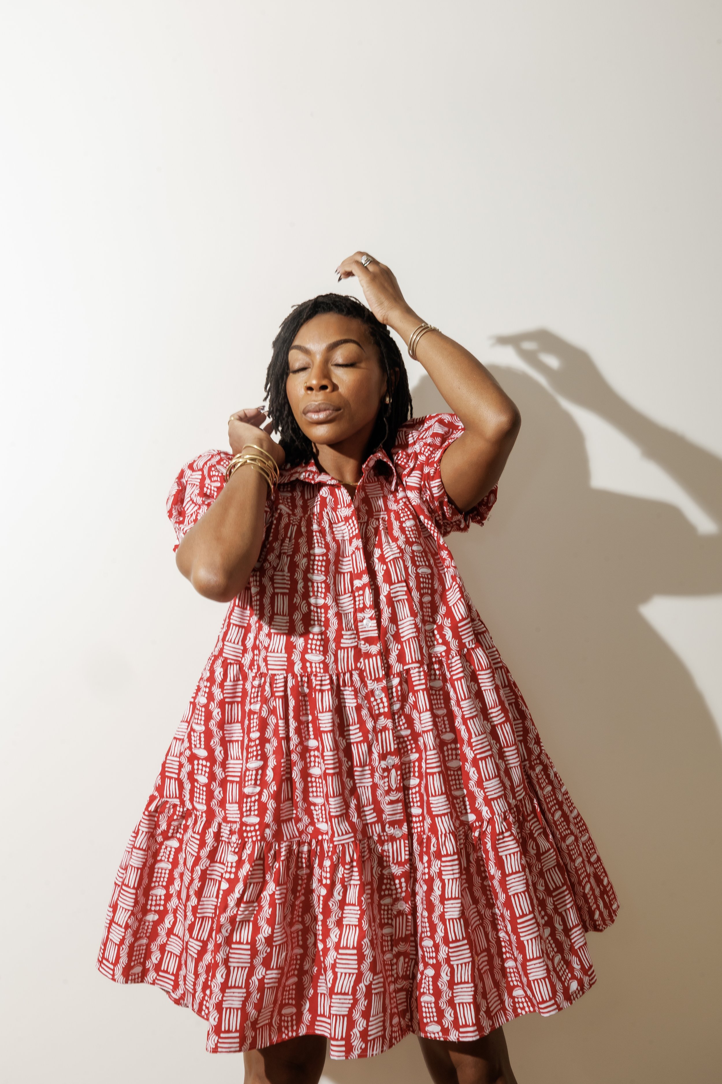 A woman with medium skin tone and closed eyes, wearing a red and white patterned dress with a midi length, accessorized with multiple gold bracelets, a ring, and earrings, standing against a plain white wall with her left hand touching her head and h