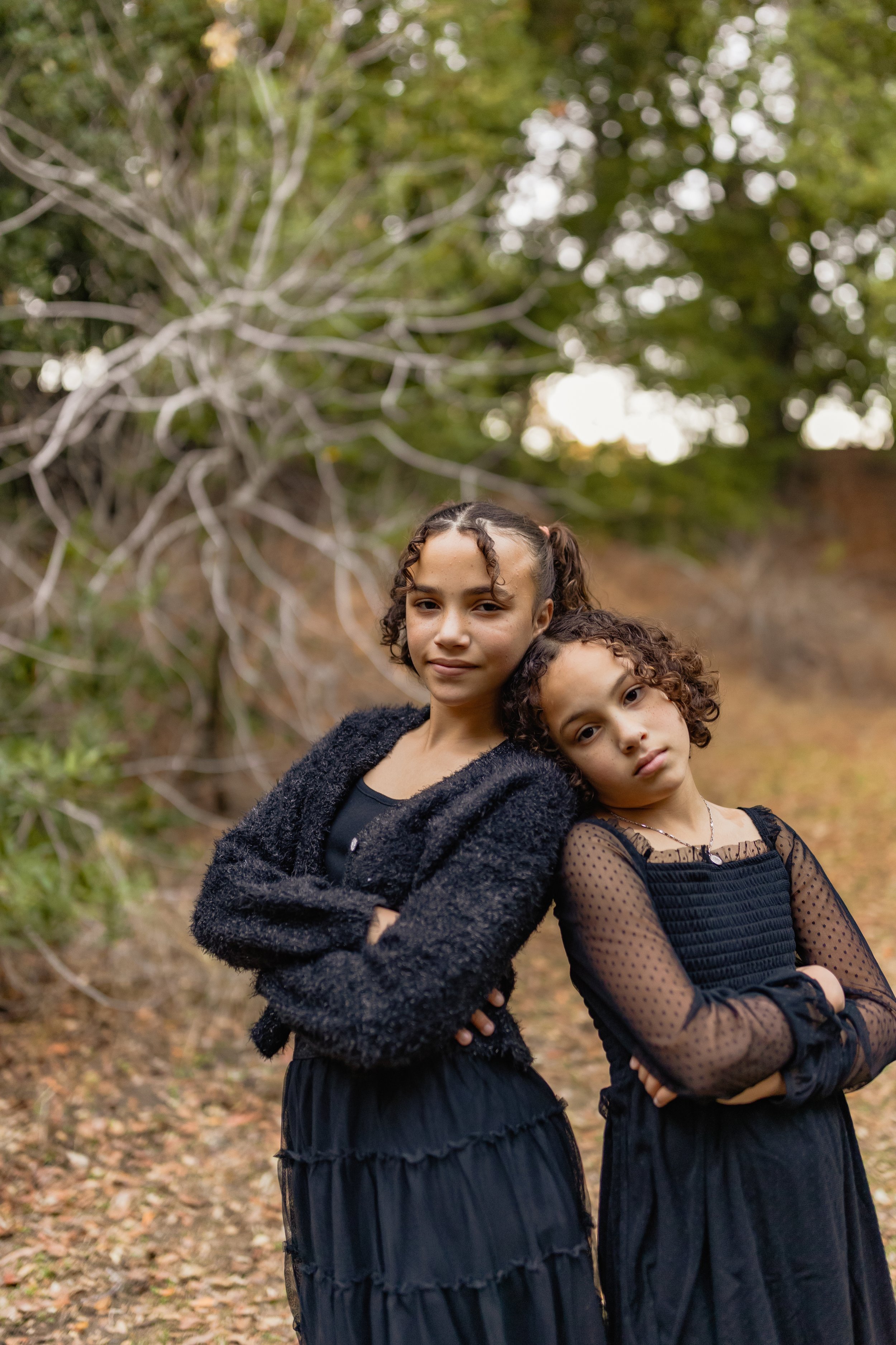 Two young girls standing outdoors on a fall day, both with arms crossed, with trees and fallen leaves in the background.
