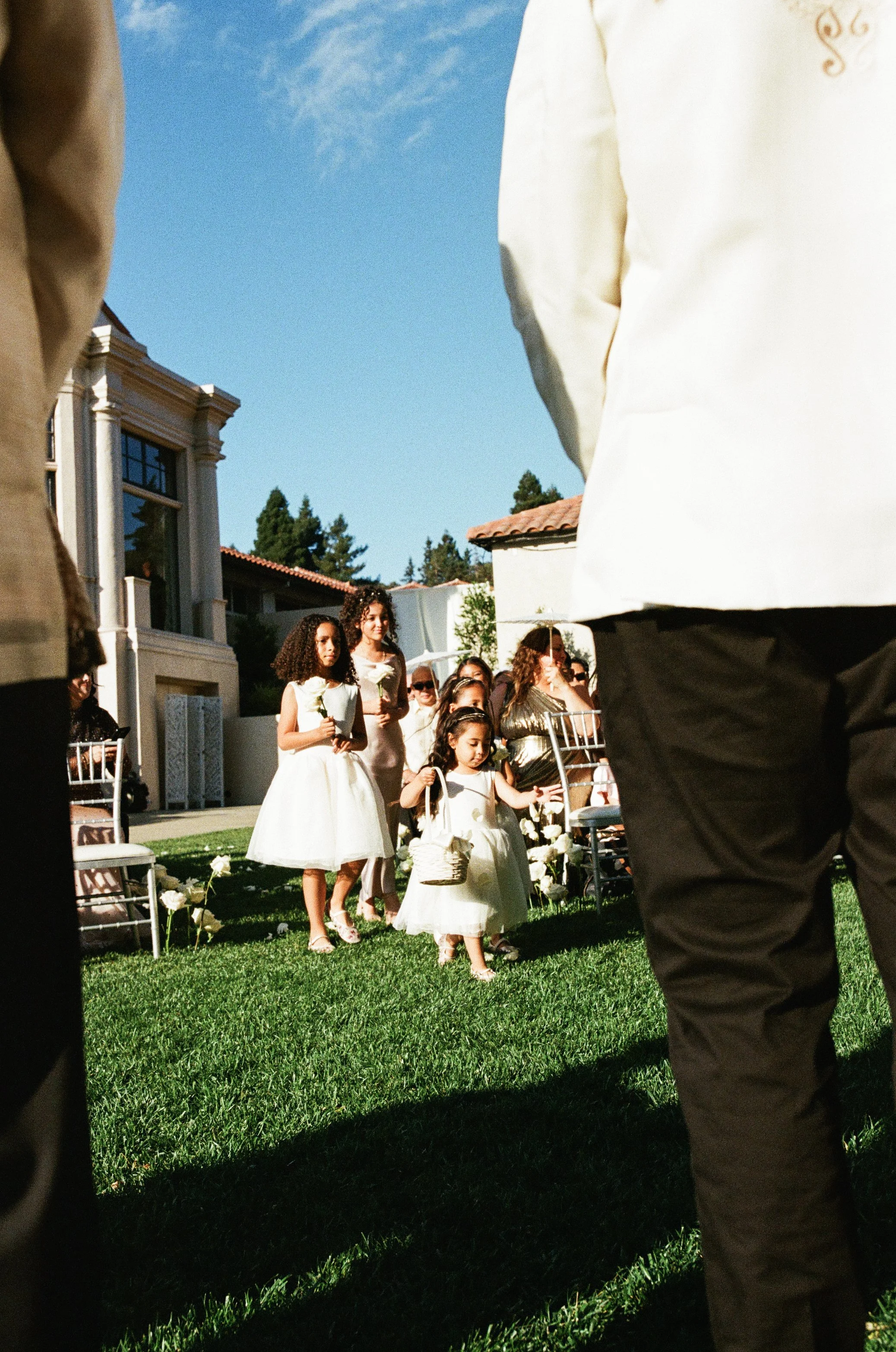 Children and adults standing in line outdoors, dressed in formal attire, likely for a wedding or special event, with a clear blue sky and residential buildings in the background.