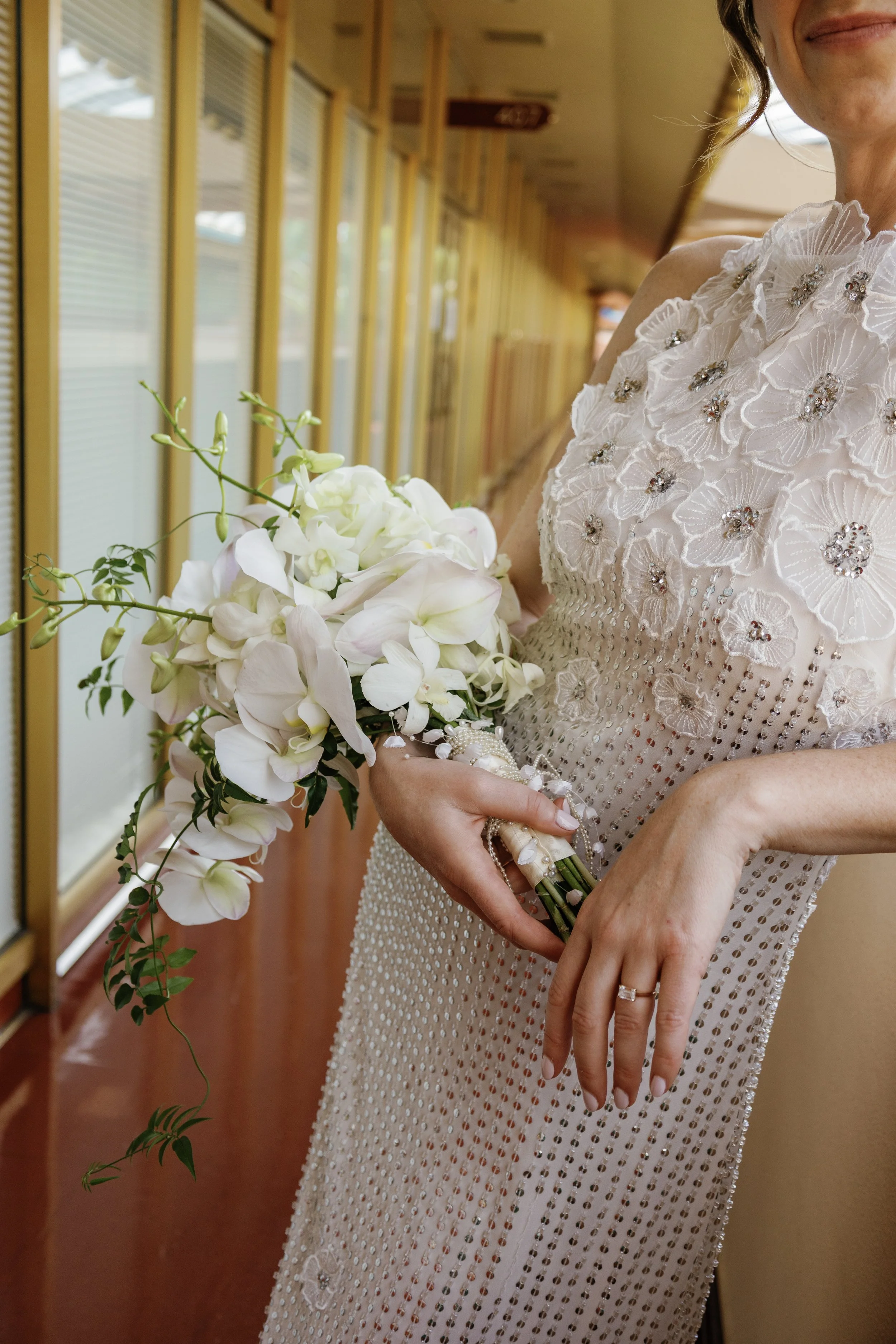 A woman in a white dress with embellishments holding a bouquet of white flowers, standing in a hallway with wooden walls and windows.