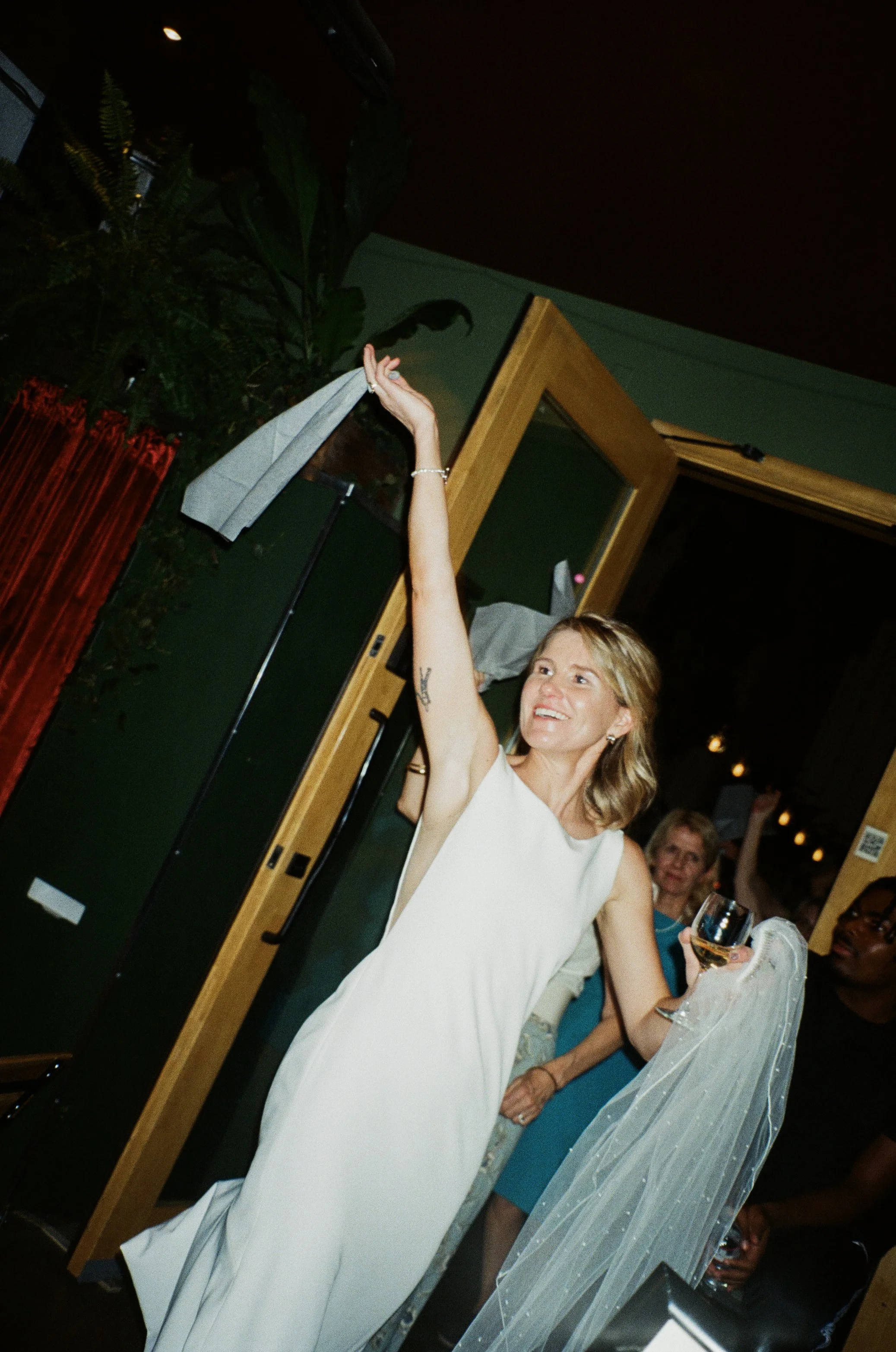 A woman in a white dress raising her hand at a party, holding a glass of white wine, with other people in the background.