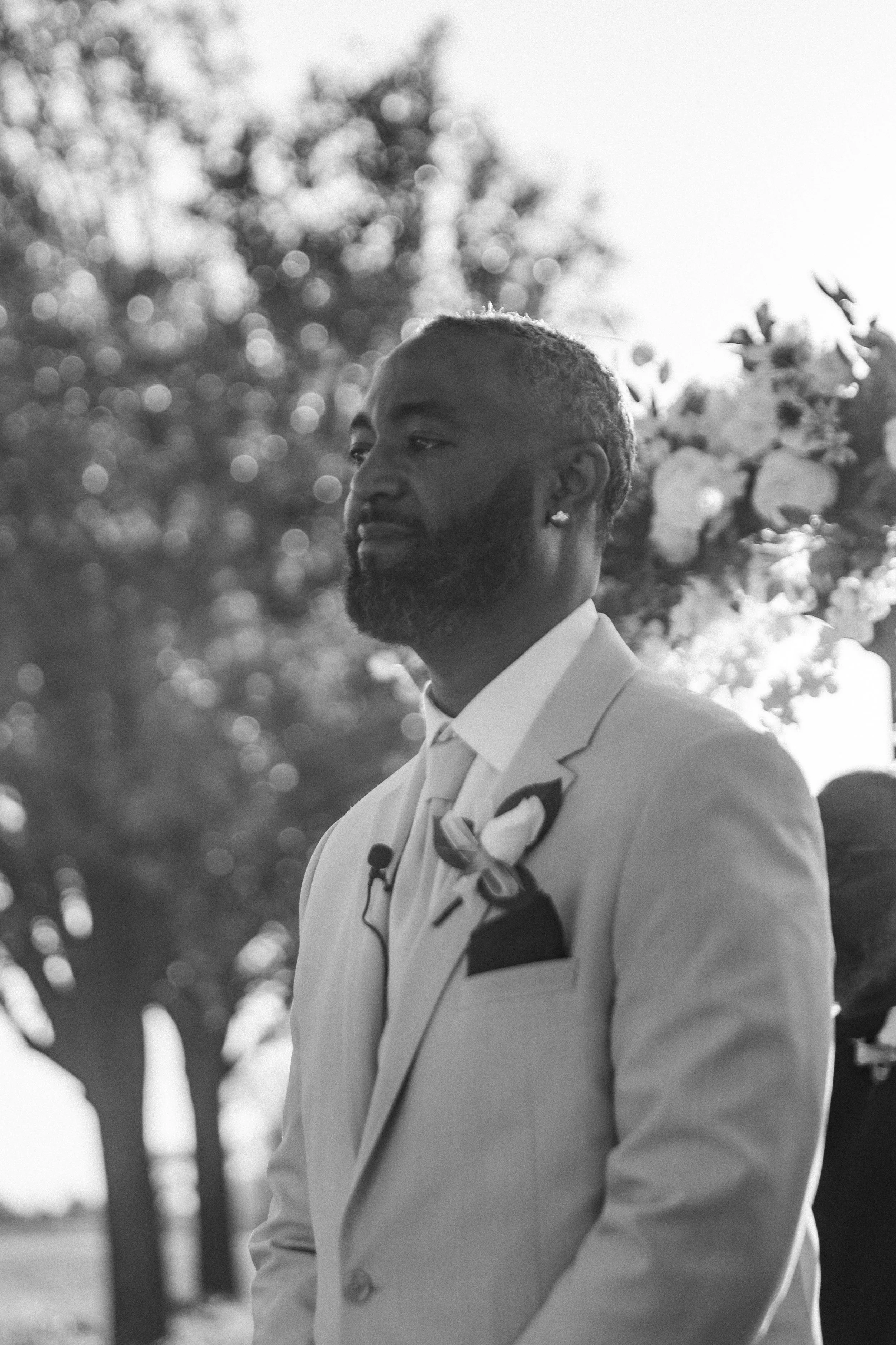 A Black man with a beard and short hair wearing a light-colored suit, white dress shirt, and boutonniere, standing outdoors with a blurred background of trees and flowers, during a wedding or formal event in daytime.