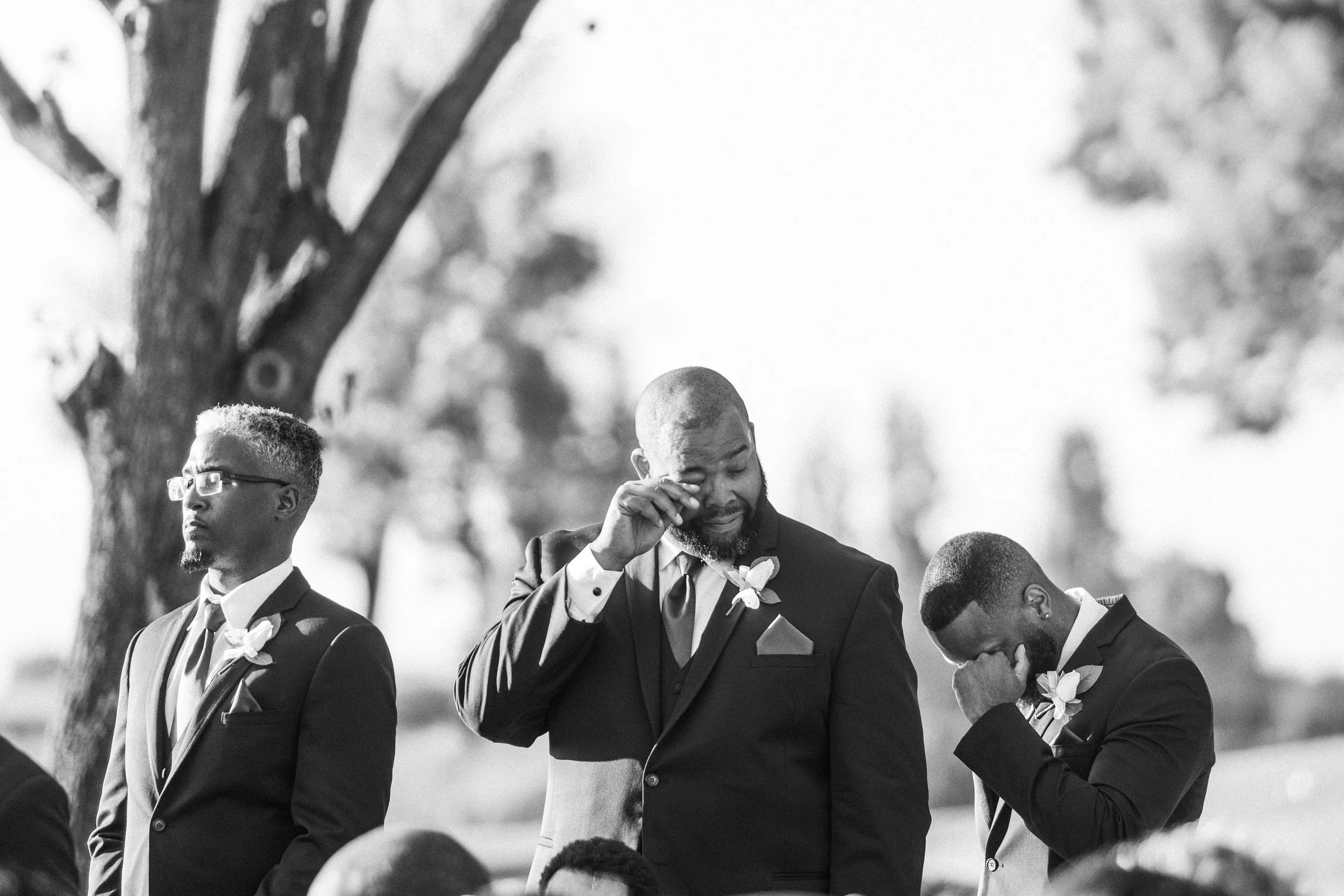 Black and white photo of a group of men dressed in suits, possibly at a wedding or formal event, with trees in the background. One man appears to be crying while others are looking down or away.