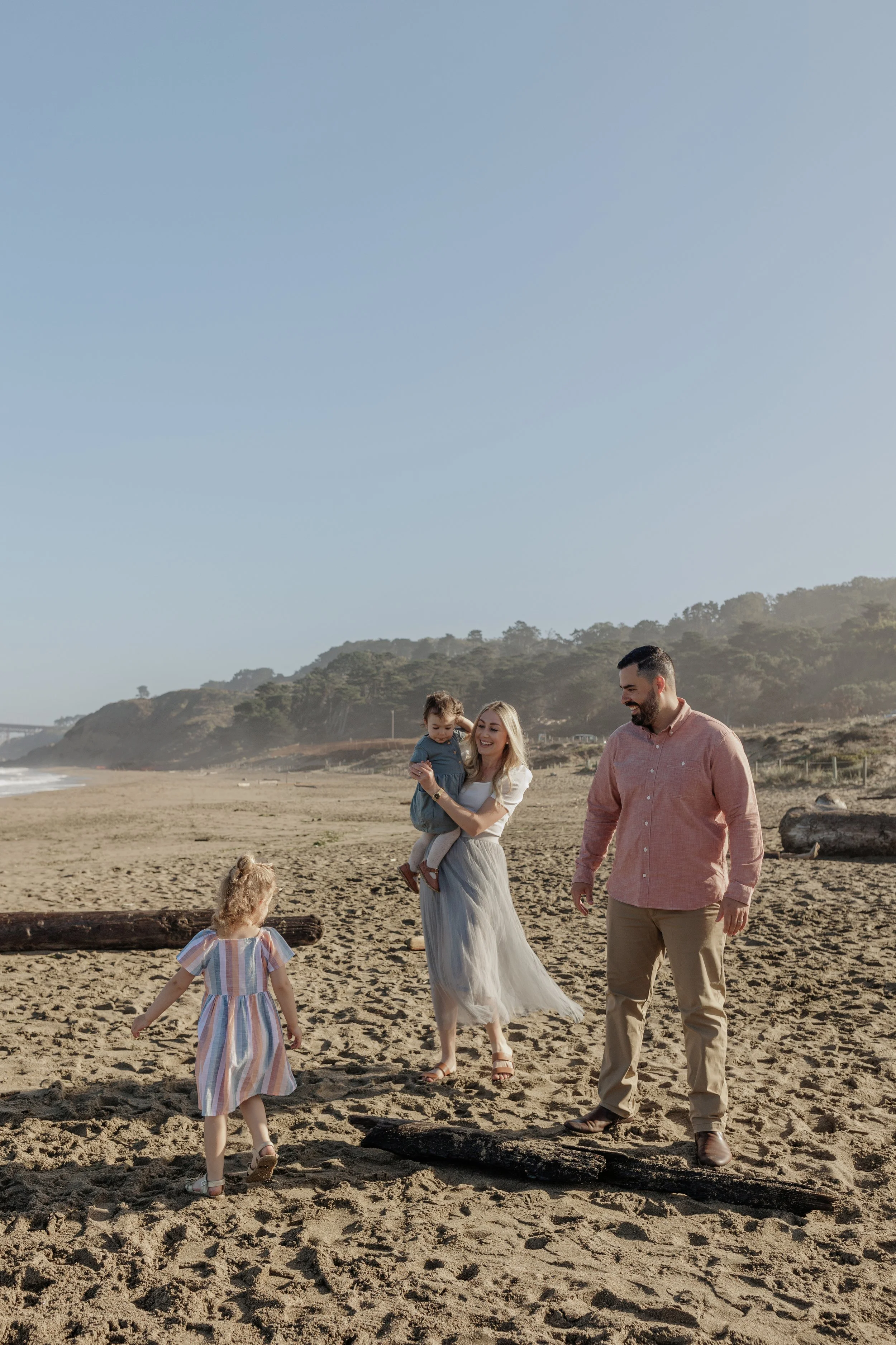 A family enjoying a walk on the beach, with a woman holding a young girl and a man walking beside them. There are two young children playing near logs on the sandy beach, with hills and a clear sky in the background.