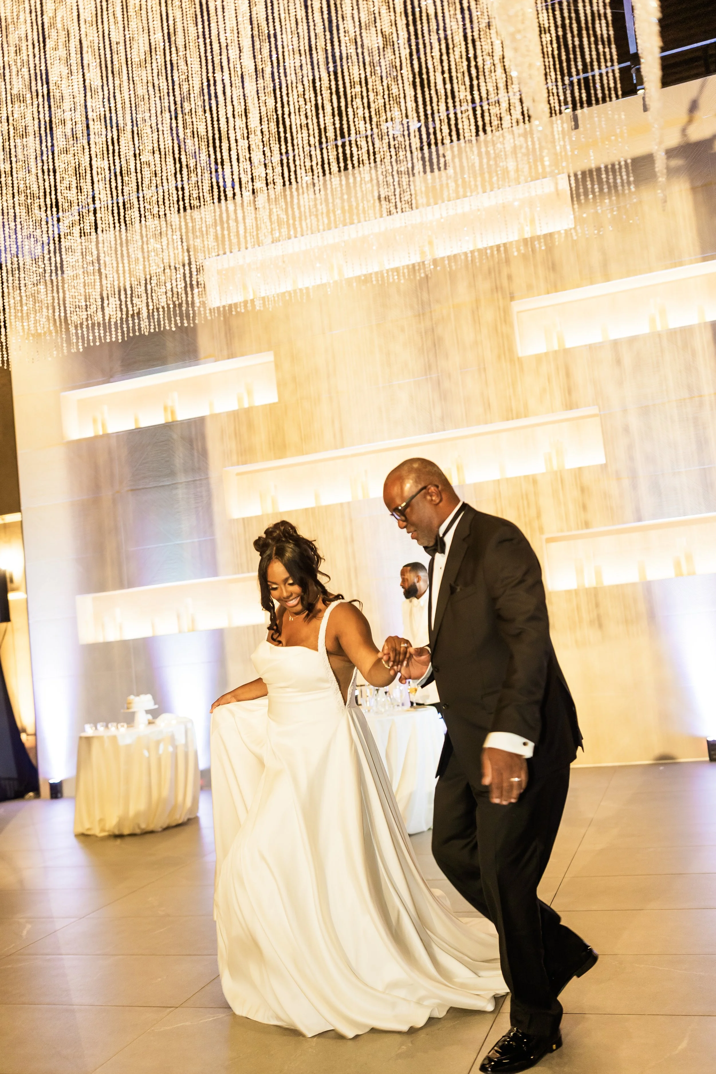 A bride and groom dancing at their wedding reception, with the bride wearing a white wedding gown and the groom in a black tuxedo, in an elegant venue with warm lighting and a decorative ceiling.