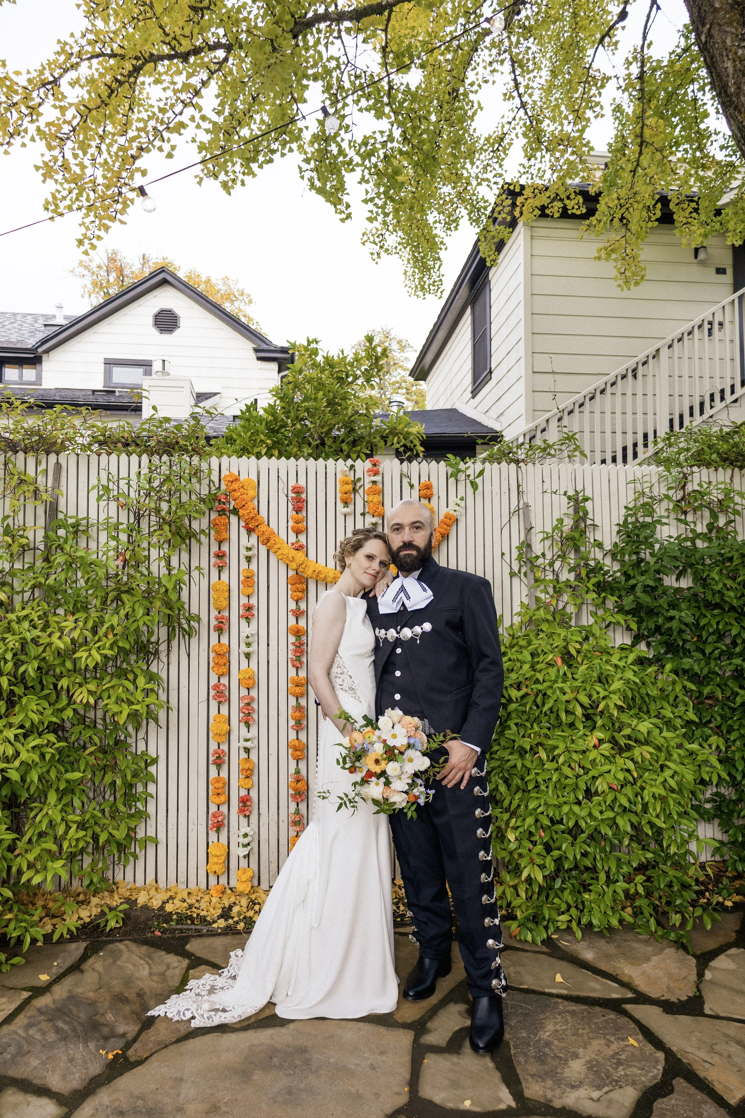 A bride and groom standing together in front of a white picket fence decorated with orange and yellow marigold garlands. The bride wears a white wedding gown with lace details and holds a bouquet of white, yellow, and peach flowers. The groom wears a
