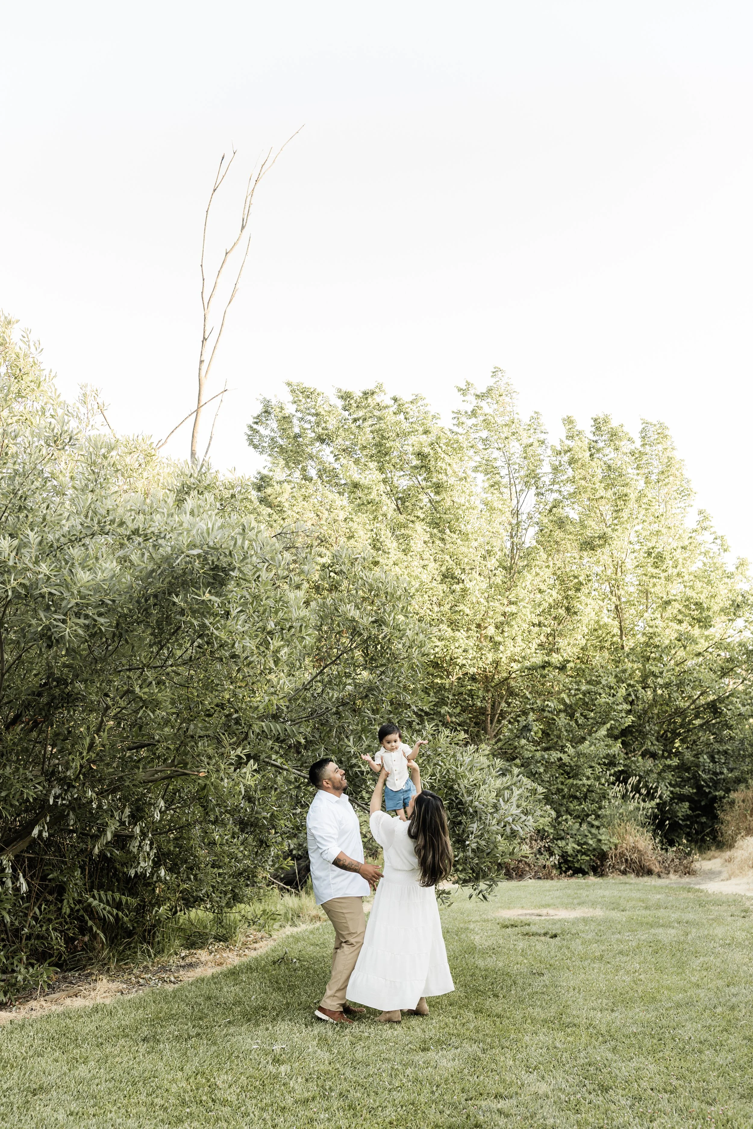 A family of three outdoors, with trees in the background and grass underfoot. A man, woman, and young child are sharing a joyful moment, with the woman lifting the child into the air.