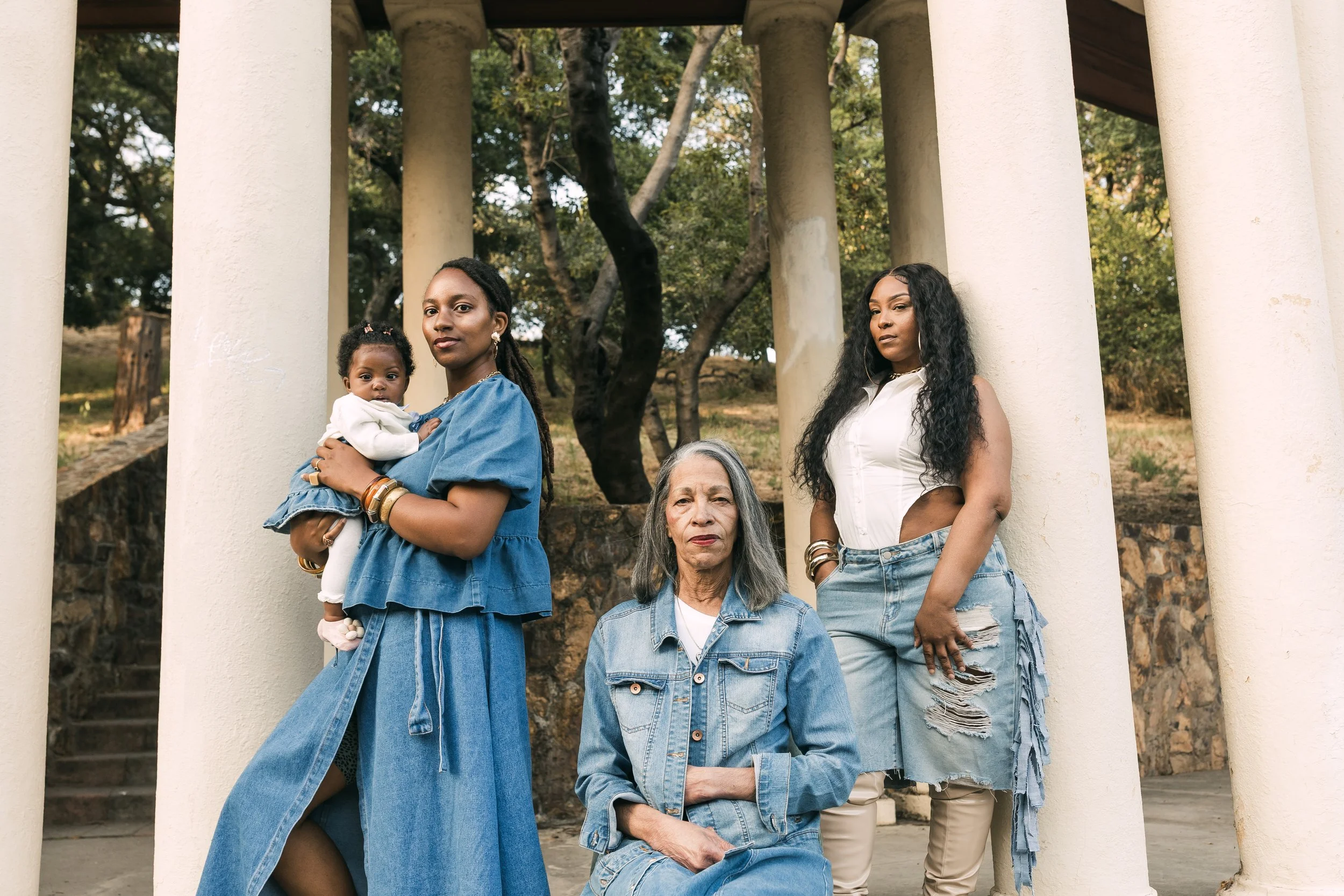 A group of five women and one young girl posing outdoors under an arched structure with columns, trees in the background, and a stone wall. The women have diverse hairstyles and are dressed in casual denim and white clothing.