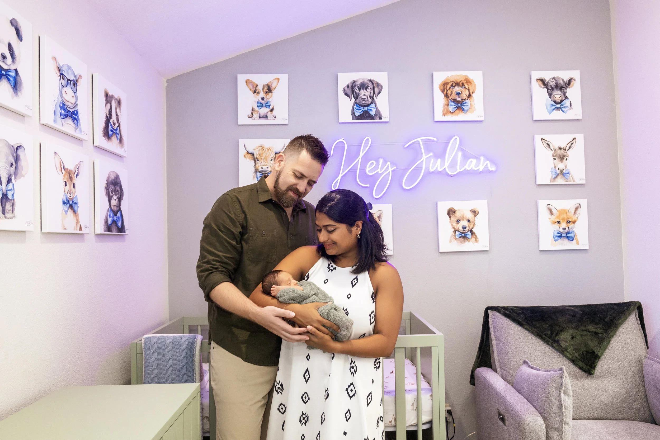 A couple is holding a newborn baby in a room decorated with dog and animal portraits wearing blue bow ties. They are smiling and looking at the baby, with a neon sign reading 'Hey Julian' in the background.