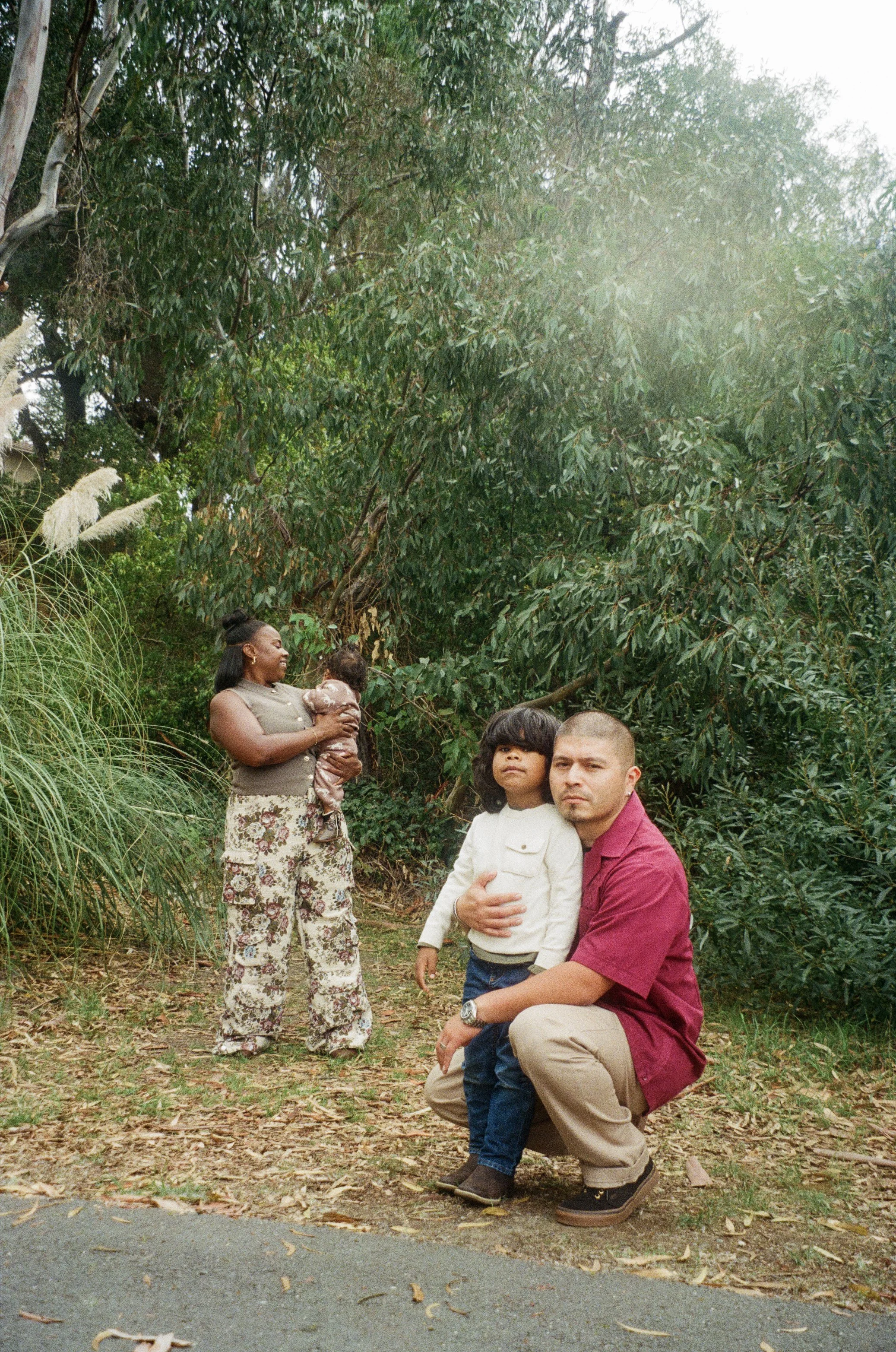 A group of people standing outdoors on a dirt path surrounded by dense green trees and grass. A woman is holding a child, and a man, kneeling in front, is holding a young boy. The group appears to be enjoying a day in nature.