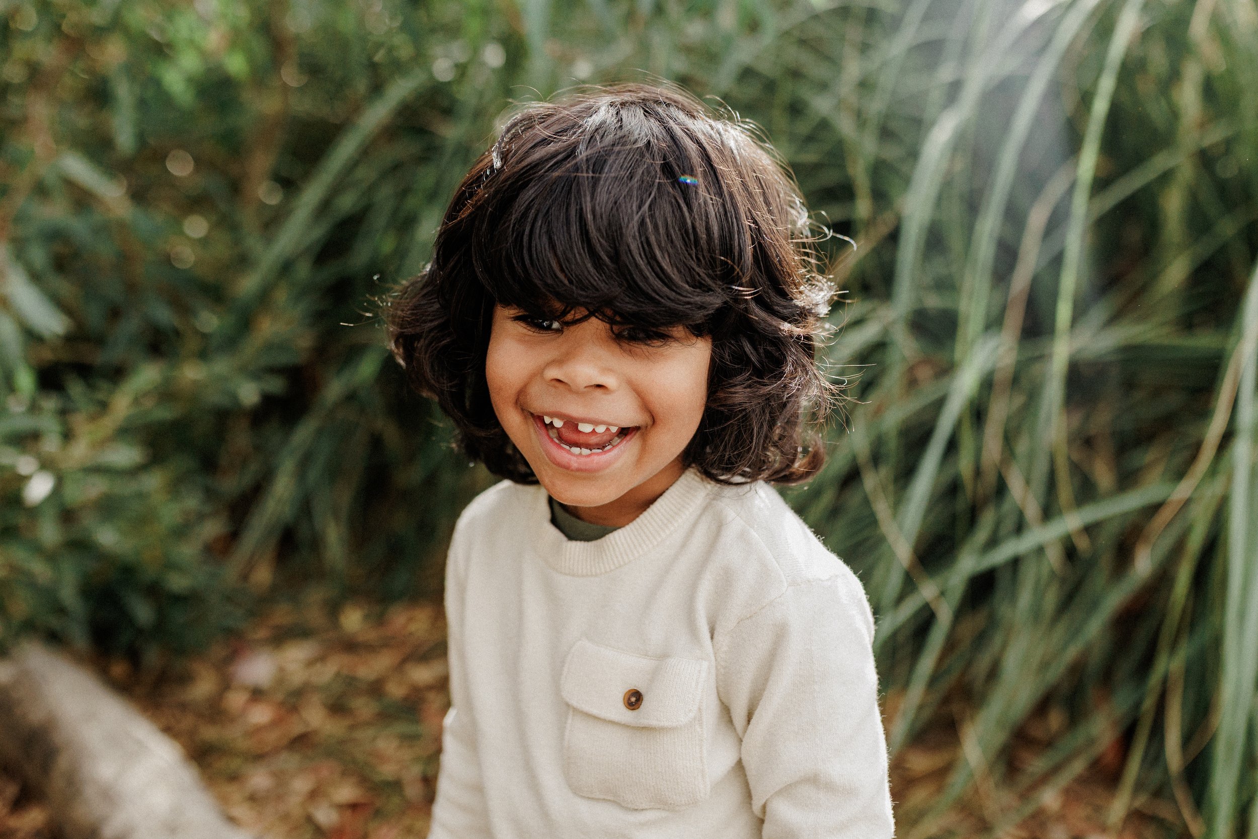 Young boy with curly dark hair and missing front teeth smiling outdoors in front of green plants.