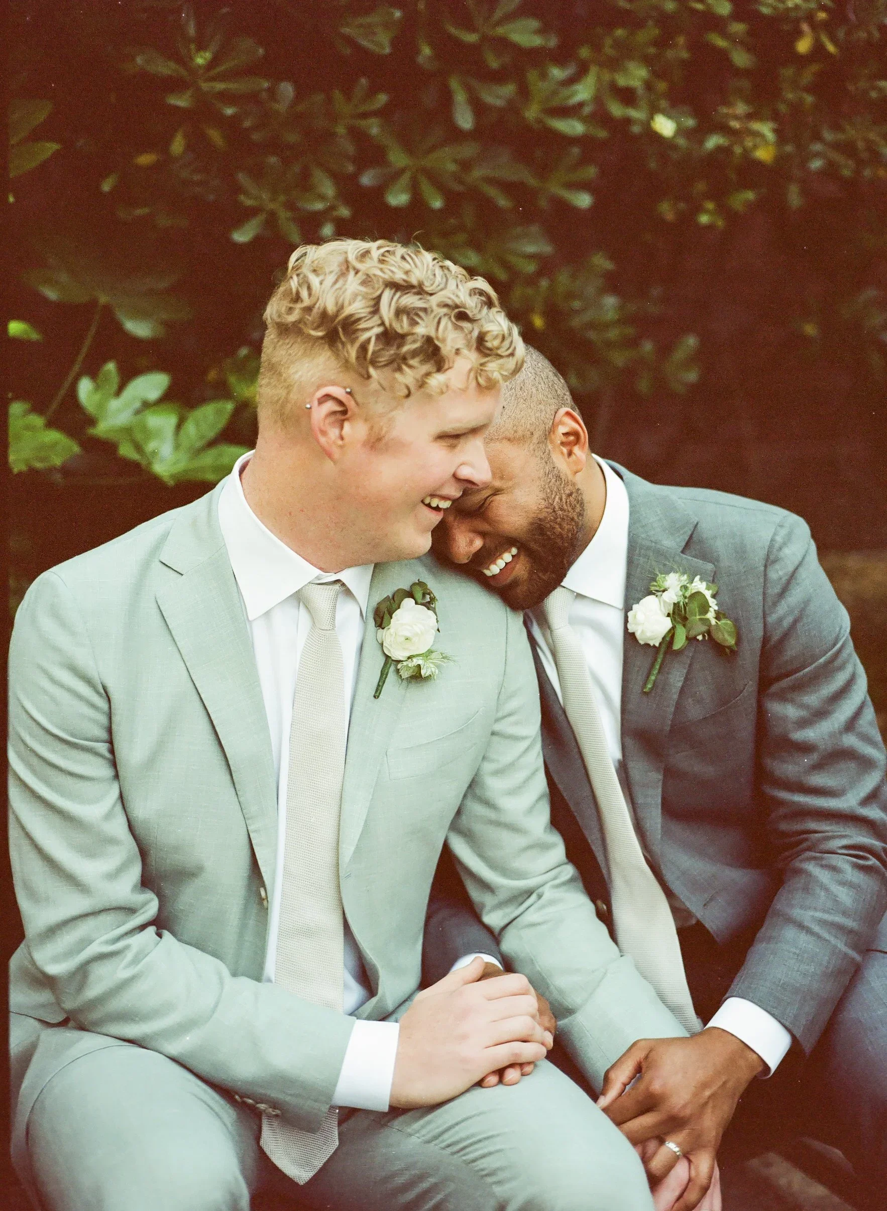 Two men dressed in suits are sitting closely together, smiling and sharing a moment of intimacy. They are outdoors with a background of green leafy bushes. Both have flowers pinned to their suits, indicating a wedding or formal event.