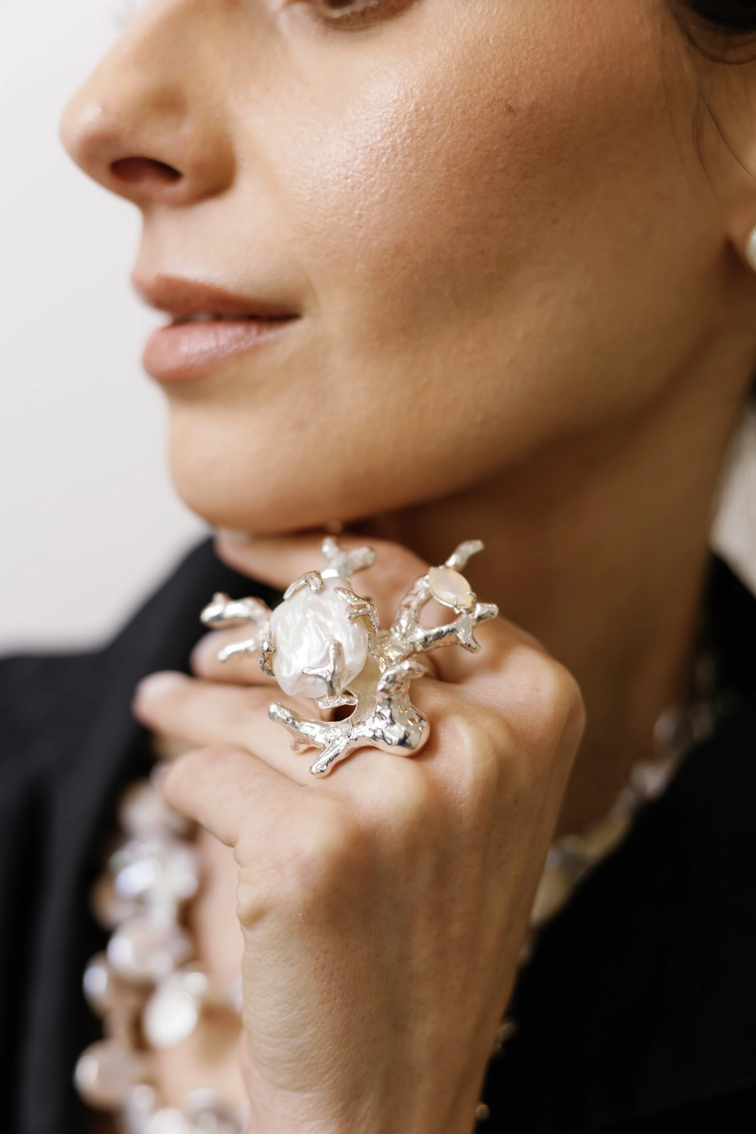Close-up of a woman wearing a large silver and pearl crab-shaped ring on her finger, and a pearl necklace, with a blurred background.
