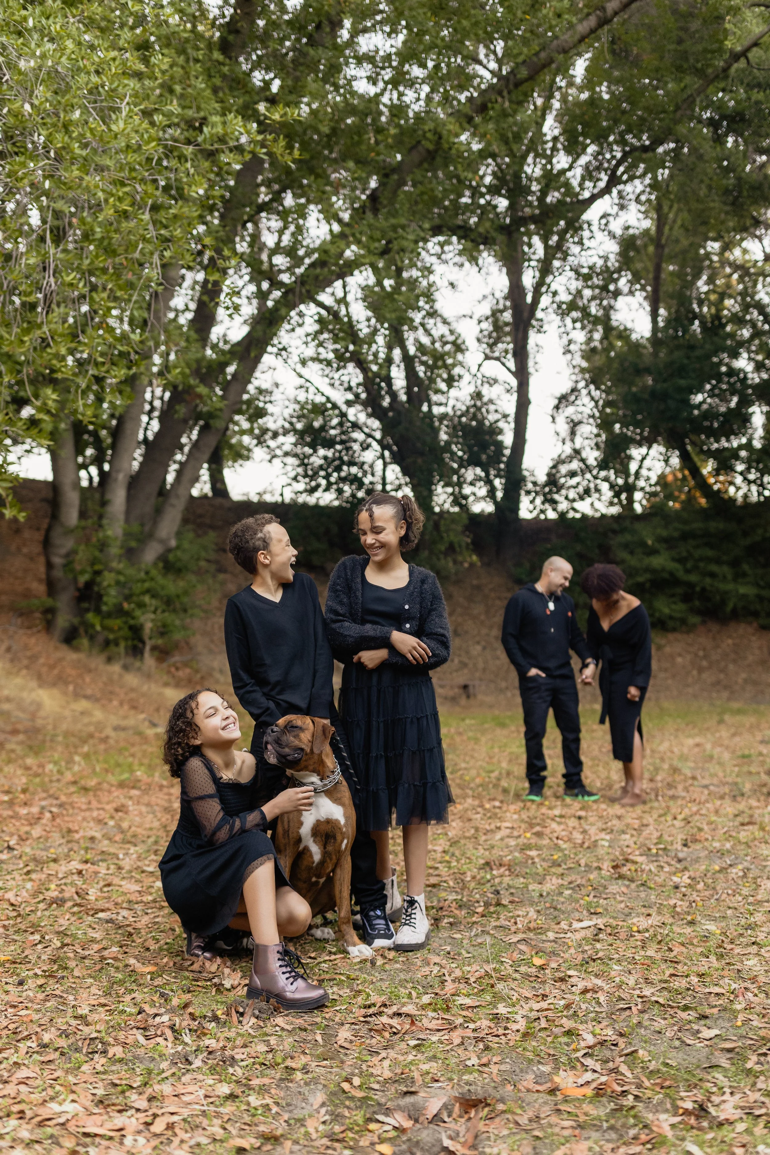 Group of people gathered outdoors on a fall day with trees and fallen leaves, including two women and a girl with a dog, smiling and talking.
