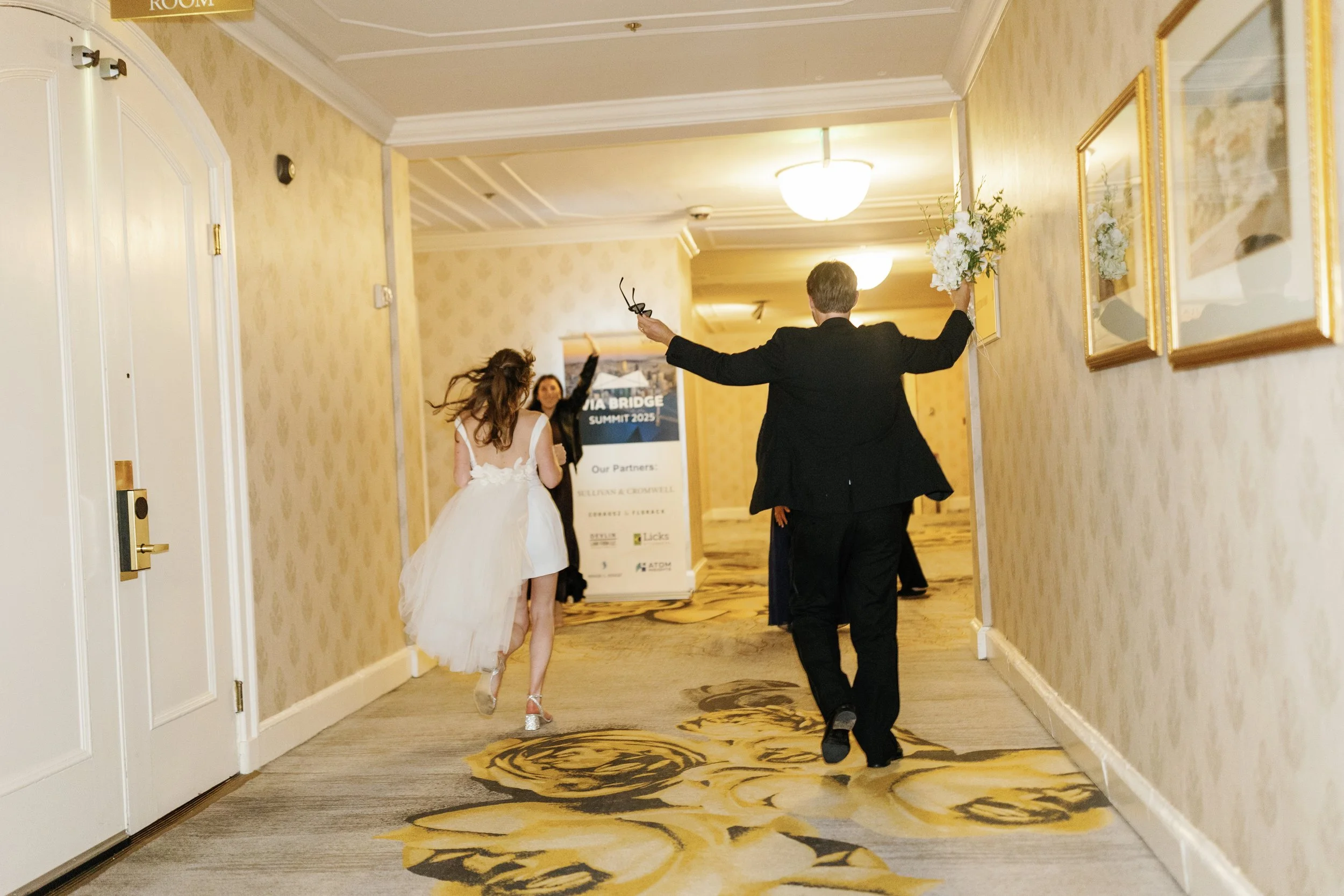 A bride in a white wedding dress running down a hotel hallway with two men, one holding a bouquet and the other holding glasses, celebrating in the background.