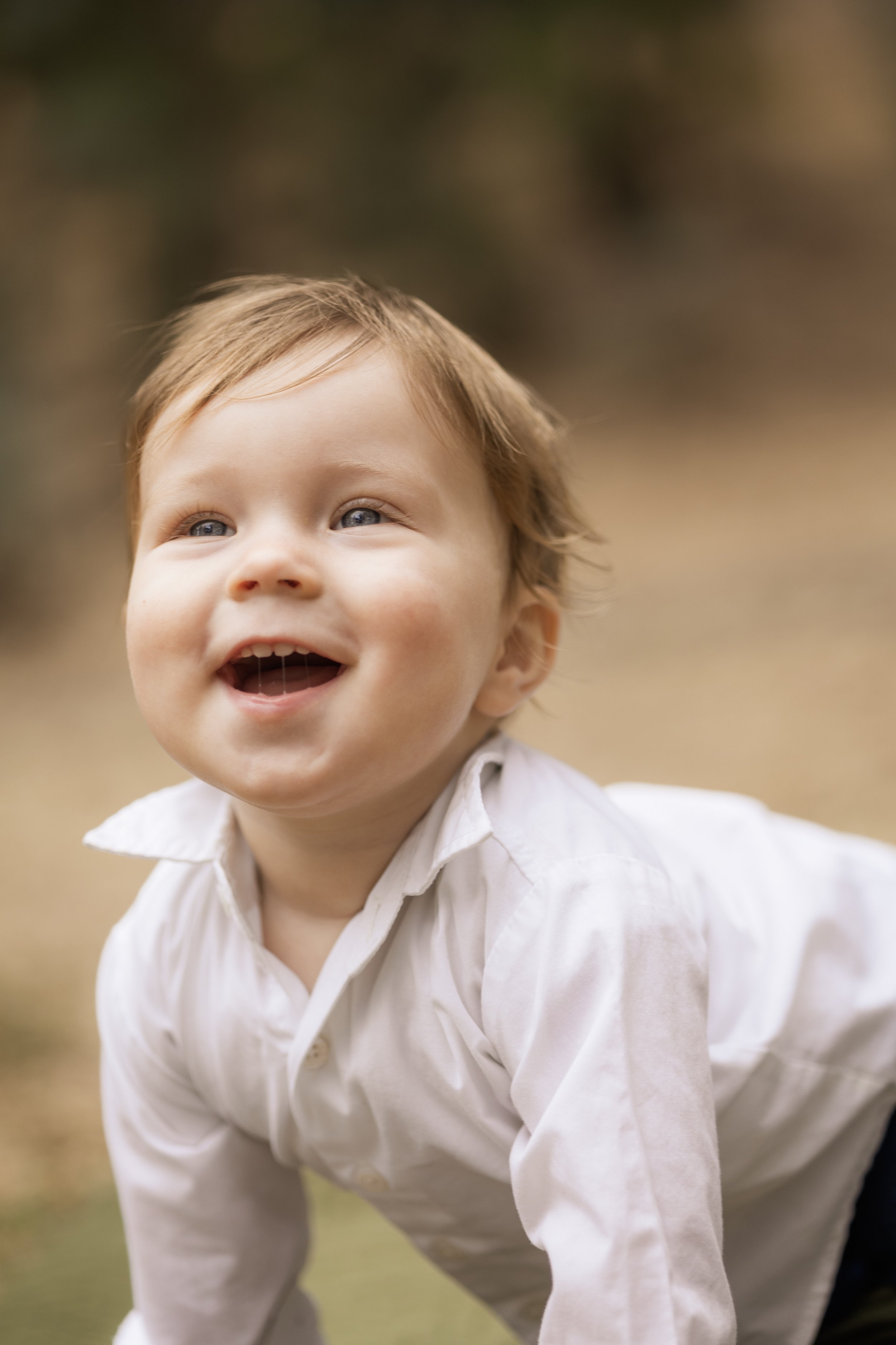 Close-up of a young child with brown hair and blue eyes, smiling outdoors in a cream-colored shirt.