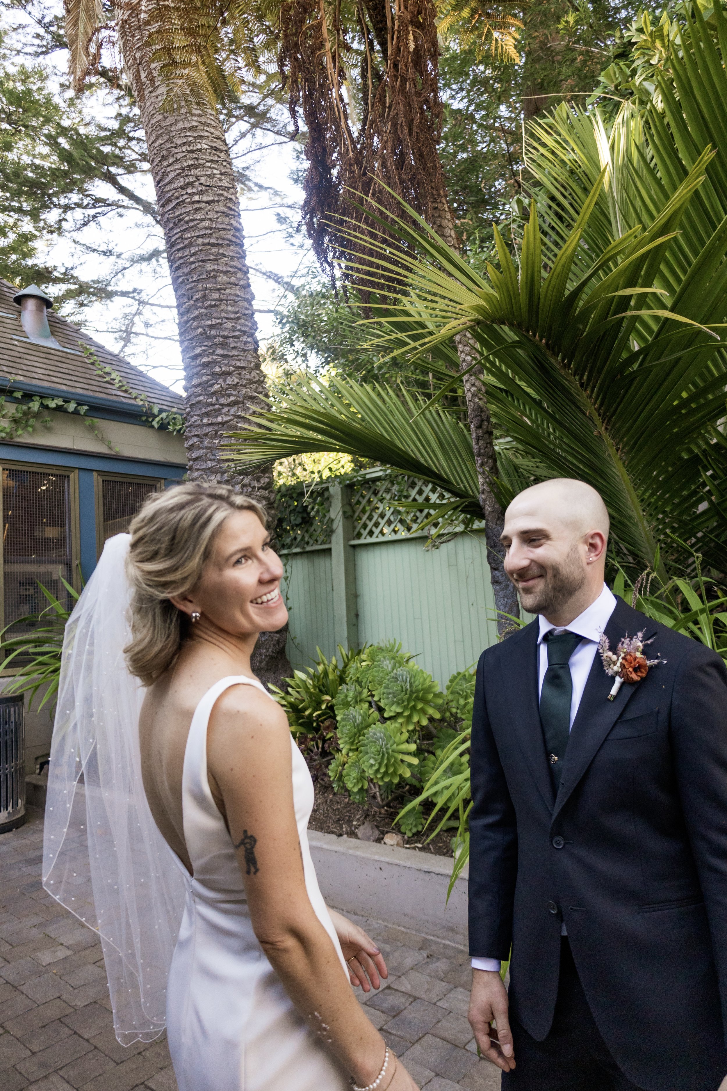 A bride with blonde hair wearing a white wedding dress and veil is smiling at a groom with a shaved head and beard, dressed in a black tuxedo with a dark green tie and boutonniere, outdoors in a garden setting with trees, plants, and a house in the b