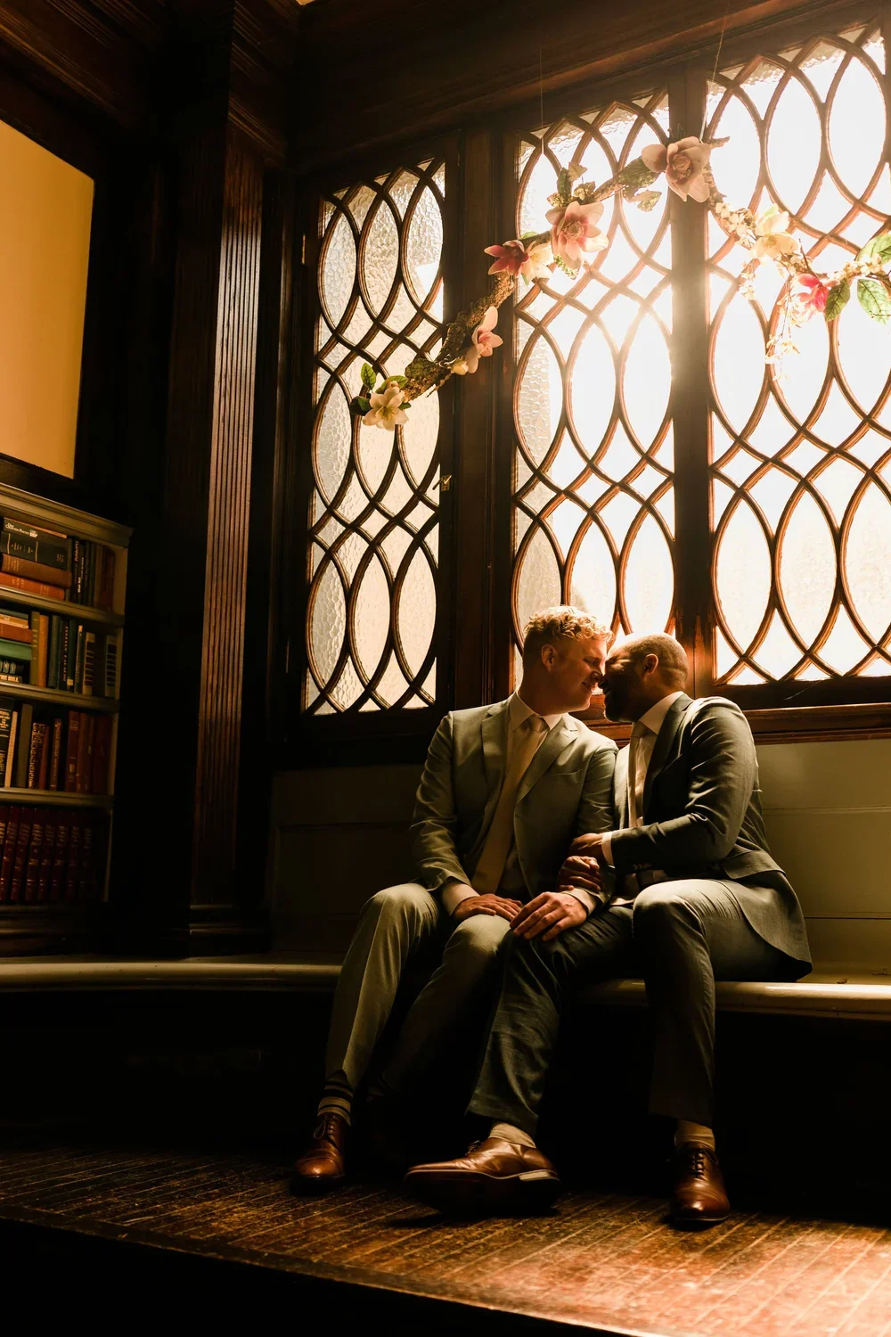 Two men in suits sitting close together on a wooden bench in front of a decorative window with a floral garland hanging across it, sharing an intimate moment in a warmly lit room with a bookshelf nearby.