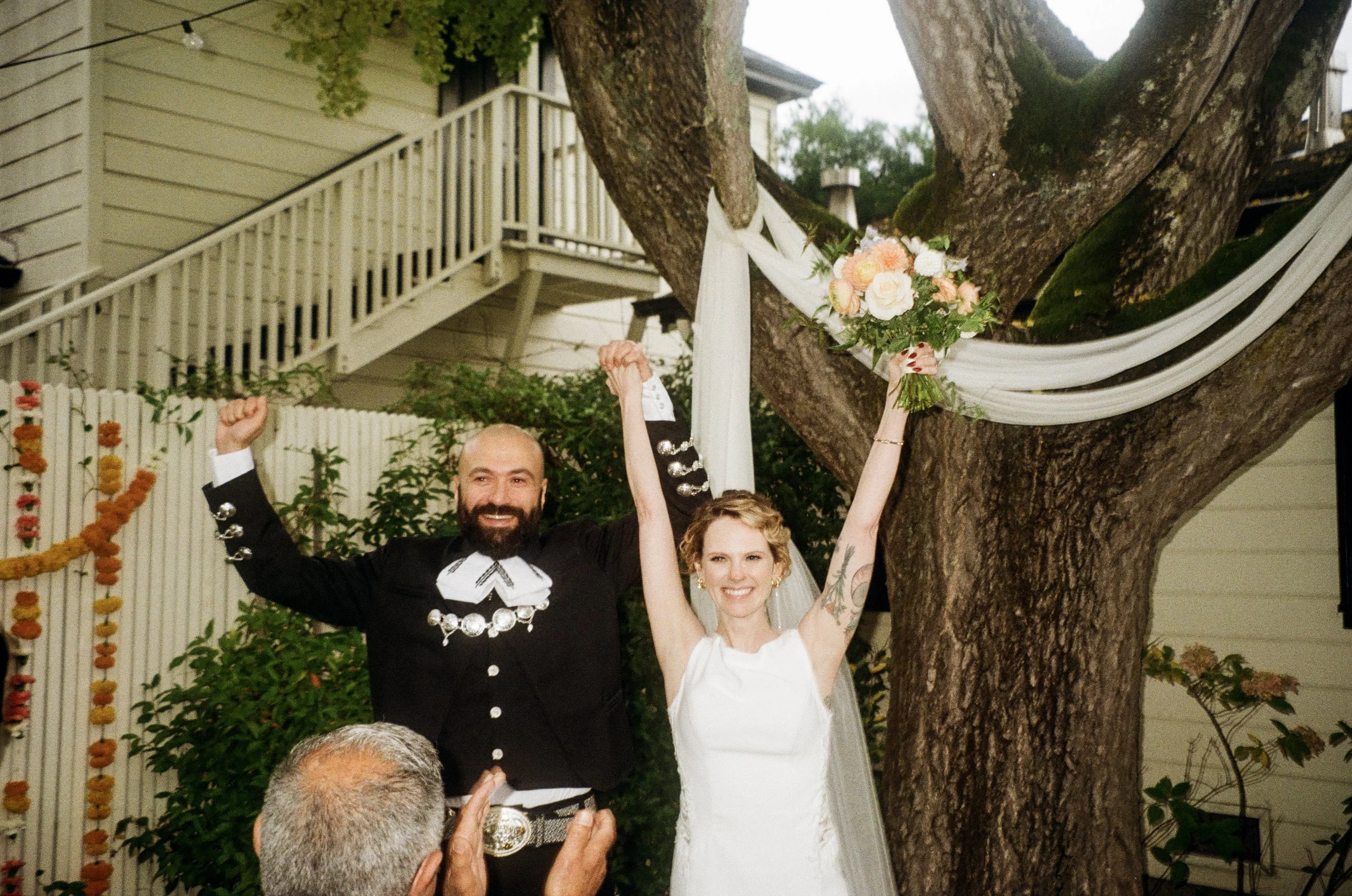 A smiling bride and groom standing under a large tree decorated with white fabric and floral arrangements at an outdoor wedding celebration.