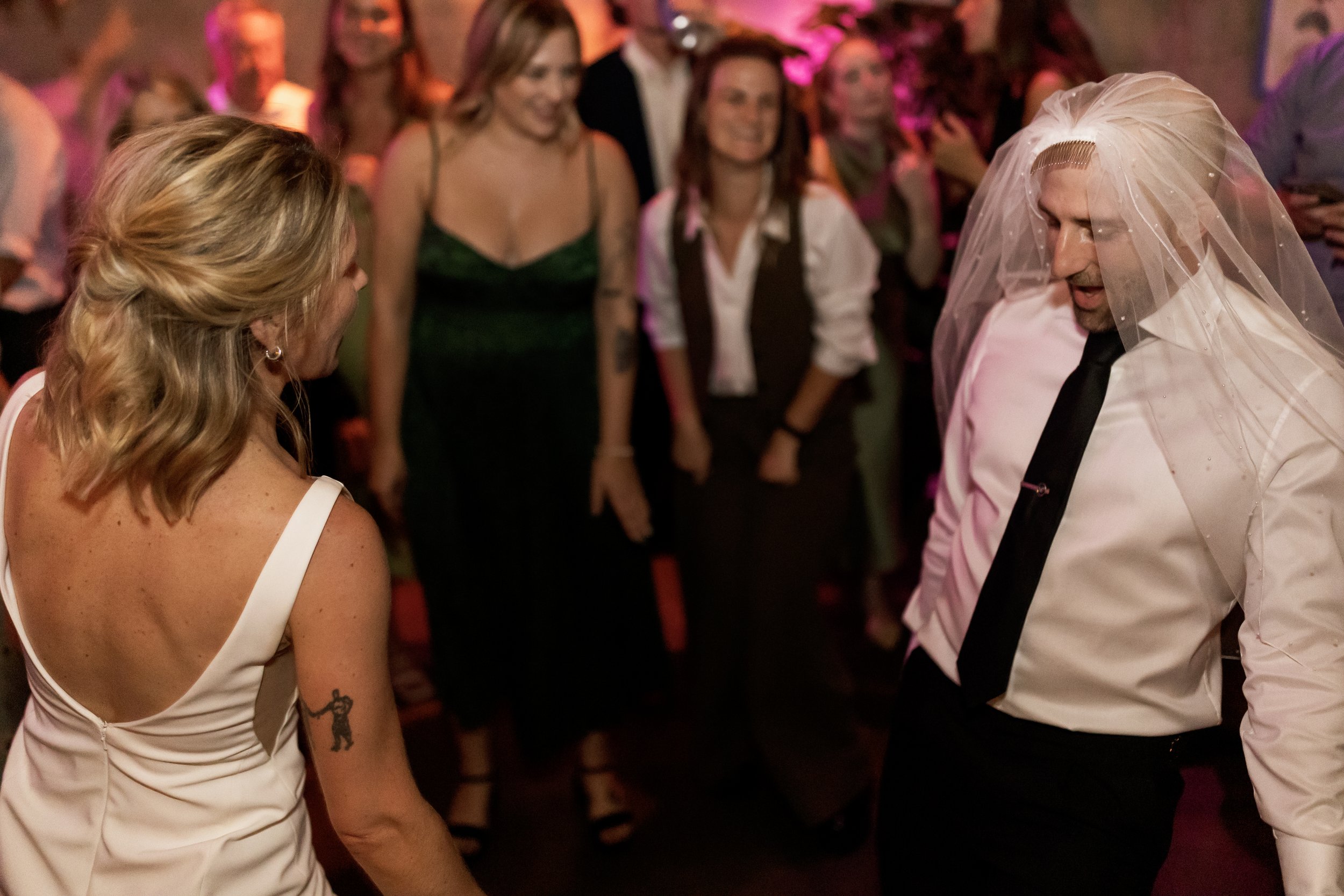 Group of people dancing and socializing at a party or celebration. The scene includes women and men, with one woman wearing a white dress and a man wearing a white shirt with a black tie and a veil, possibly dressing as a bride or for a themed event.