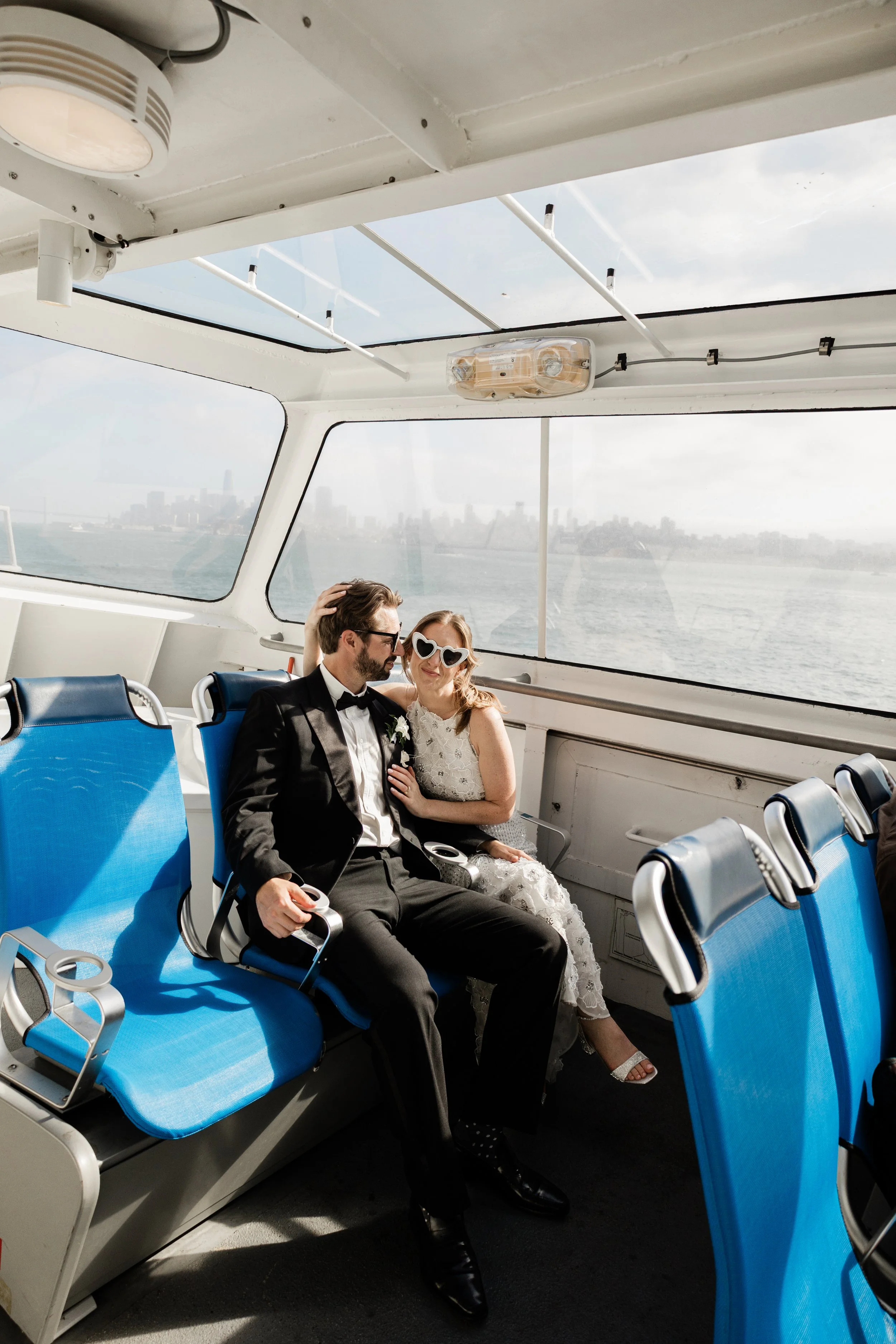 A newlywed couple sitting together on a ferry boat with blue seats, with a view of the water and city skyline in the background.