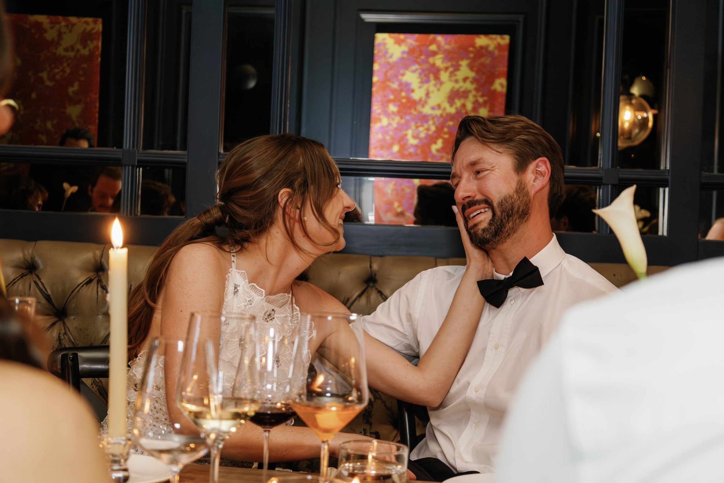 A man and woman sitting closely at a dinner table, sharing an intimate moment with the woman touching the man's face. They are dressed formally, the woman in a white dress and the man in a white shirt with a black bow tie. The table is decorated with