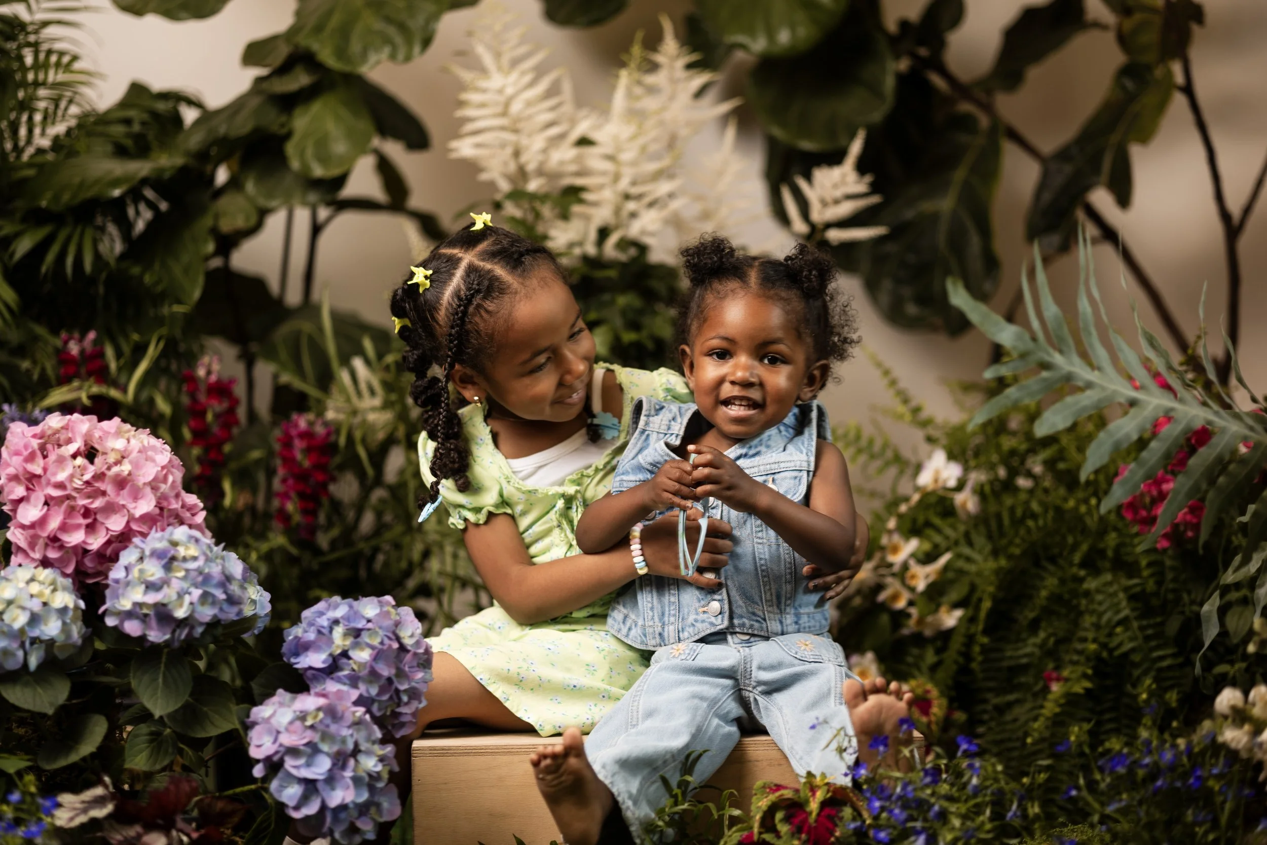 Two young girls sitting among colorful flowers and green foliage, smiling and embracing each other in a garden or floral setting.