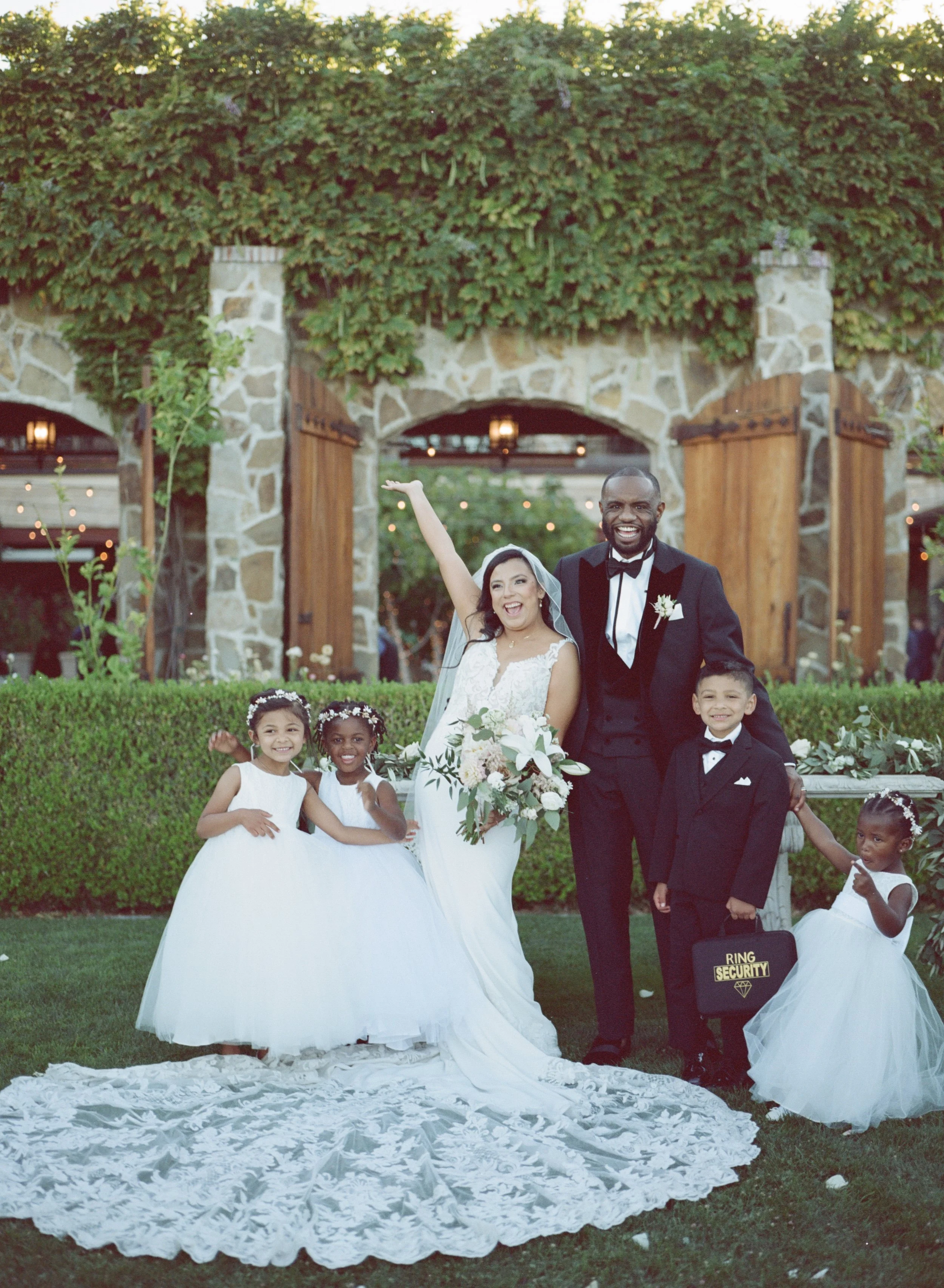 A group of wedding participants, including a bride, groom, and children, standing on a lawn in front of a stone and wood structure covered with greenery. The bride is raising her right arm and smiling, while the groom stands next to her in a tuxedo, 
