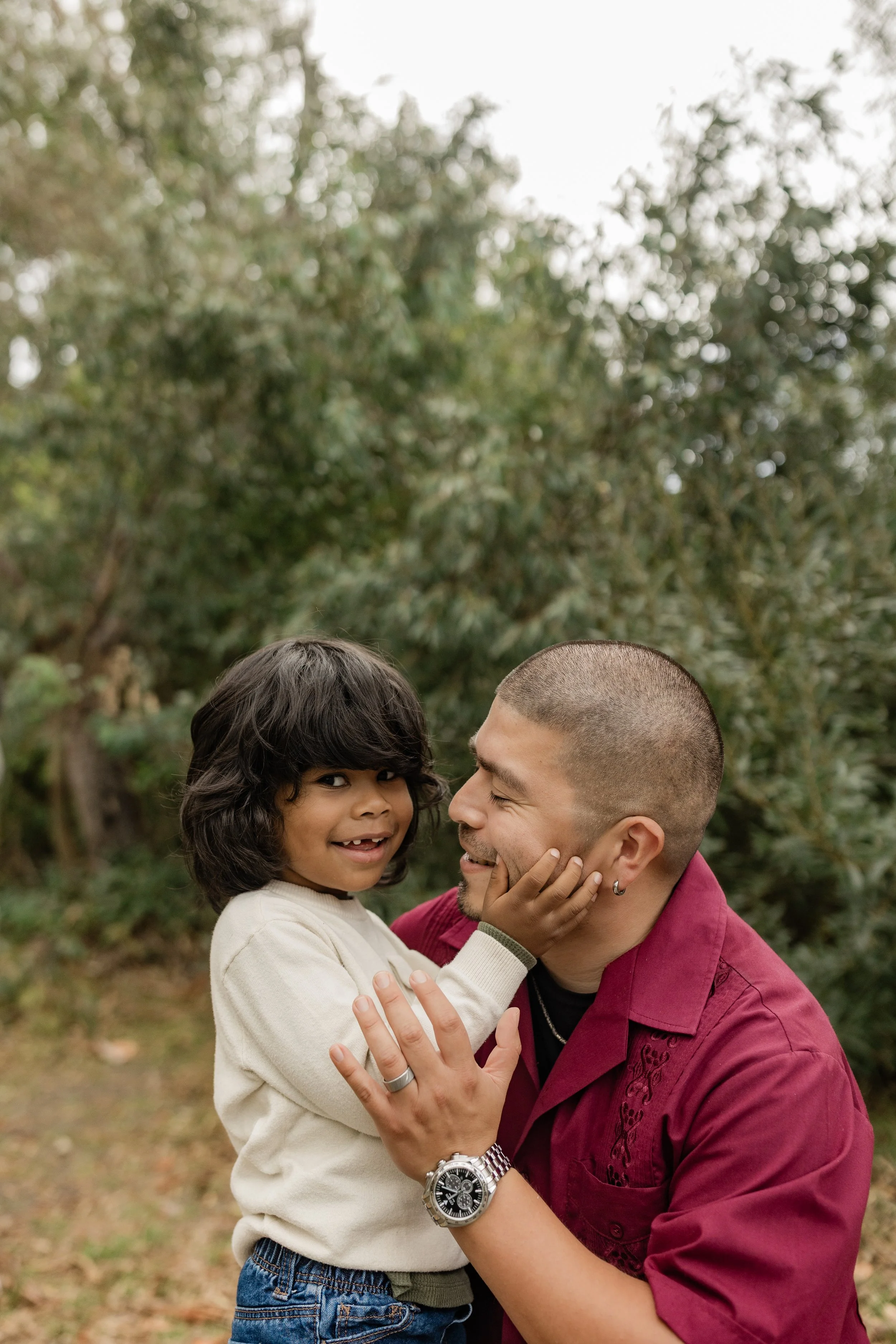 A man and a young girl are outdoors in a park or garden, surrounded by trees. The man is smiling and holding the girl by her waist, while she is smiling and touching his face with her hands. The girl has dark curly hair and the man is bald with a bea