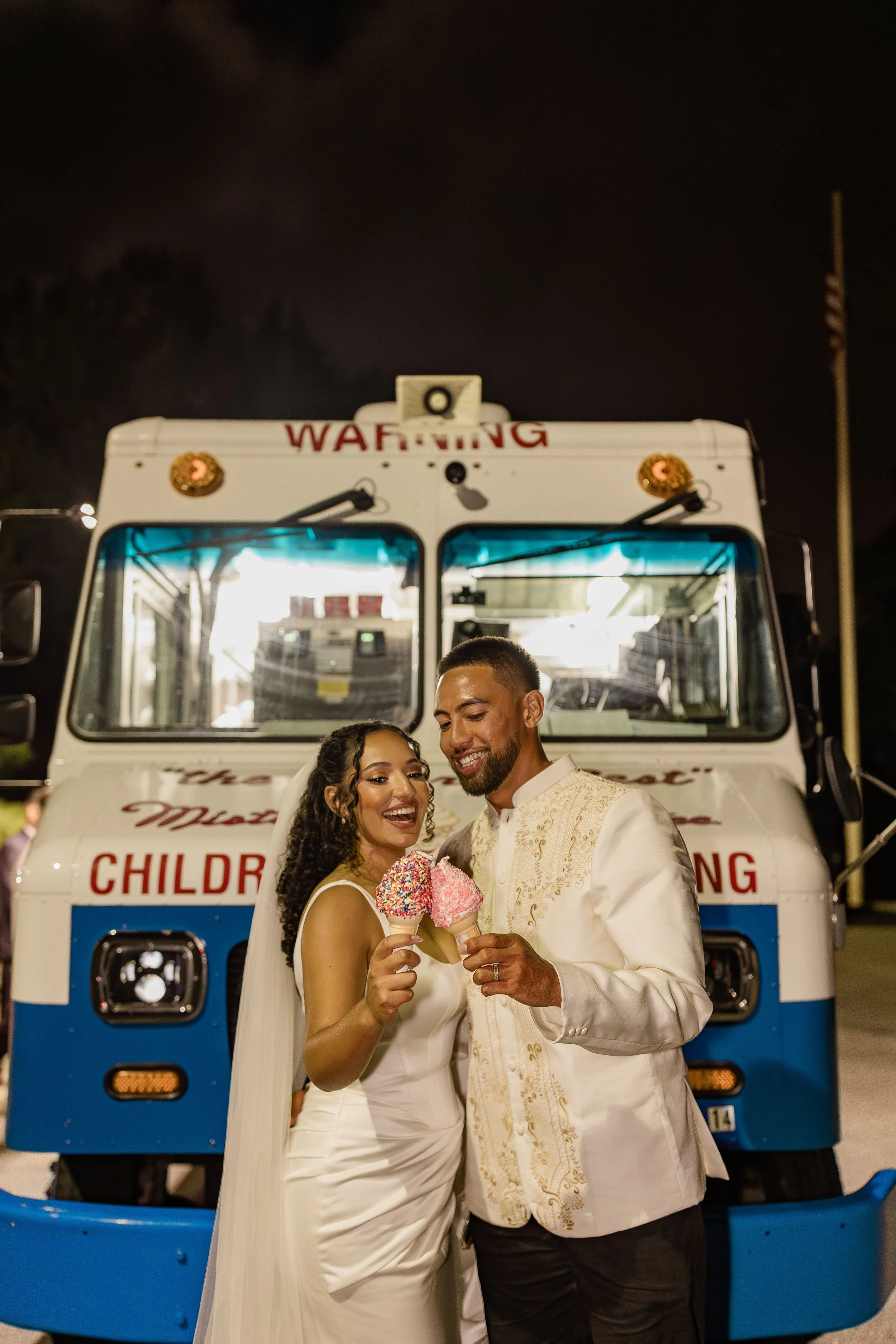 A newlywed couple standing in front of an ice cream truck at night, holding ice cream cones and smiling.