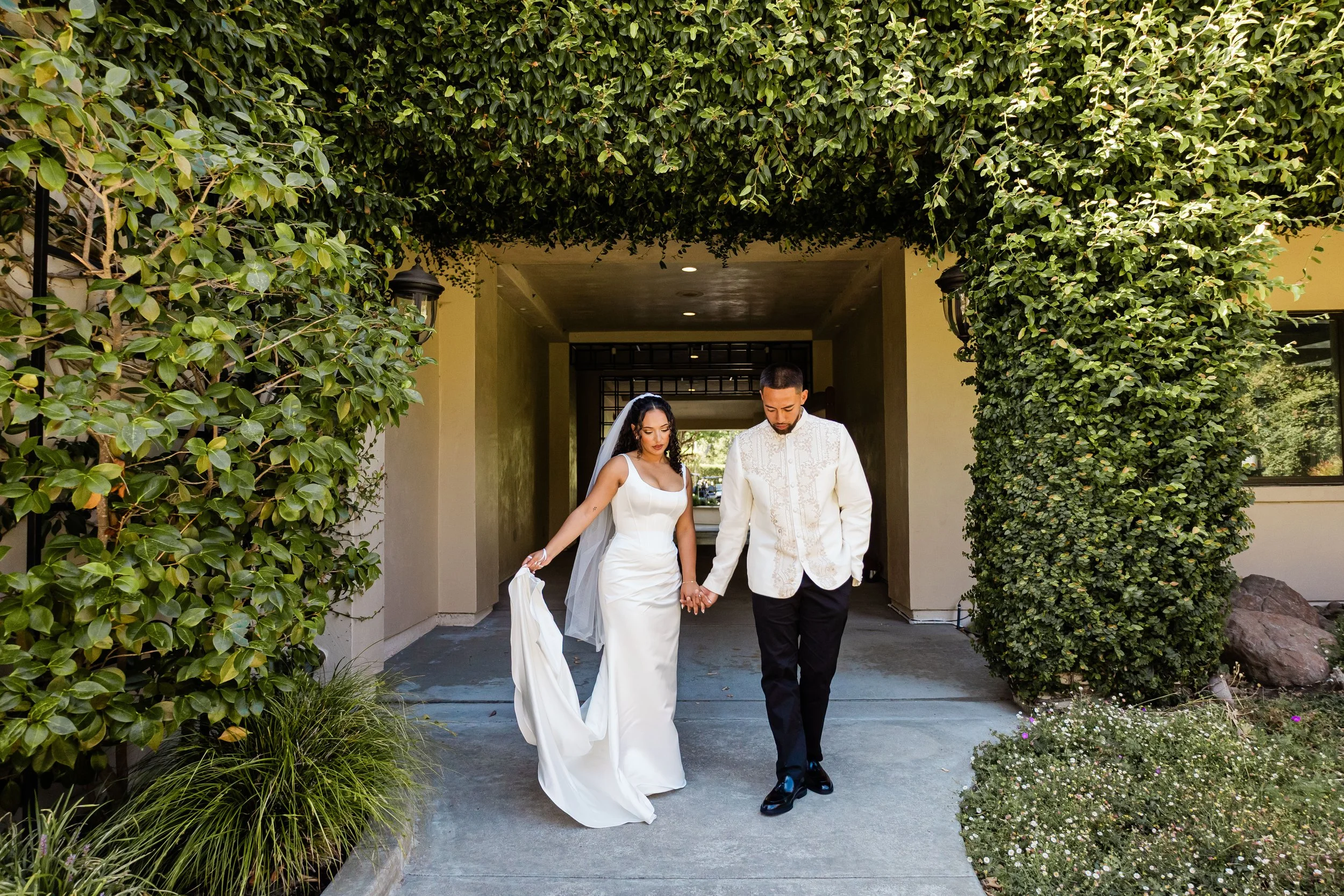 A couple dressed in wedding attire walking hand-in-hand under an archway covered with green foliage.