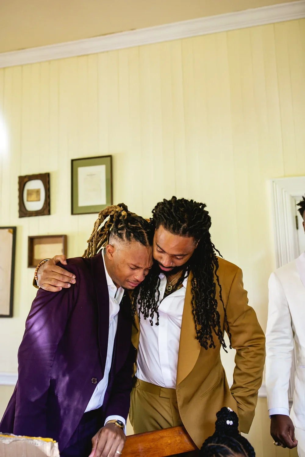 Two men are emotionally embracing each other in a church or formal setting, with one man appearing to be crying and the other comforting him. They are dressed in suits, and there are framed pictures on the wall in the background.