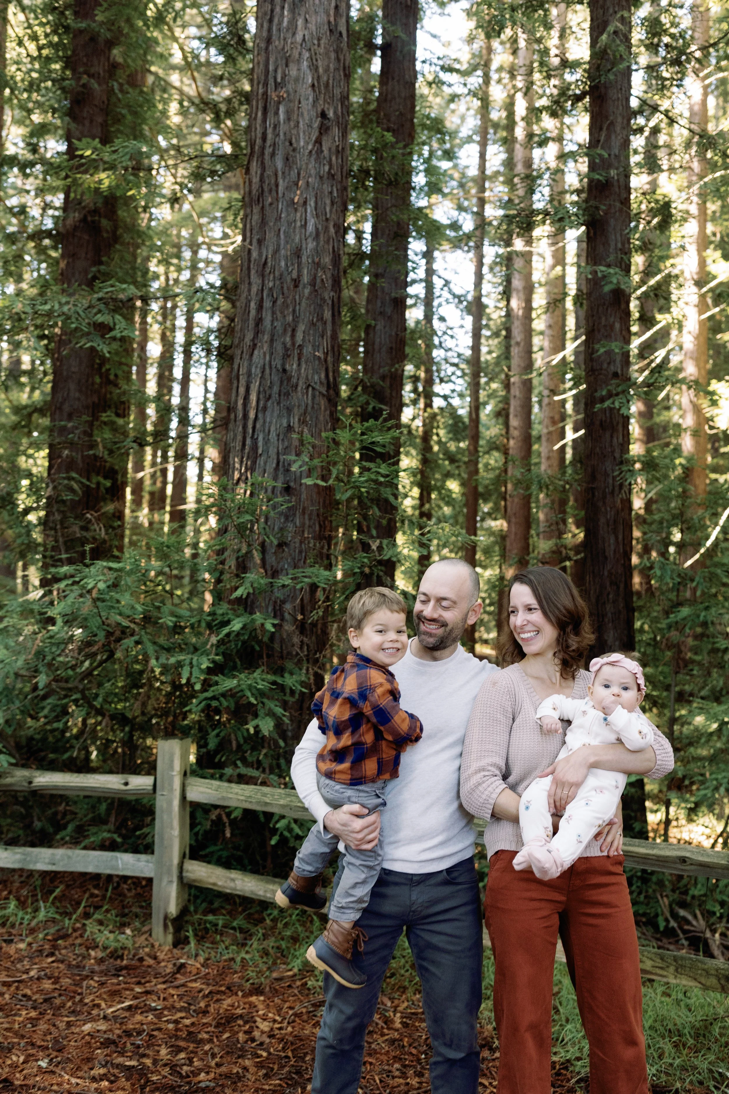 A happy family of four standing in a forest with tall trees and green foliage, with sunlight filtering through the trees.