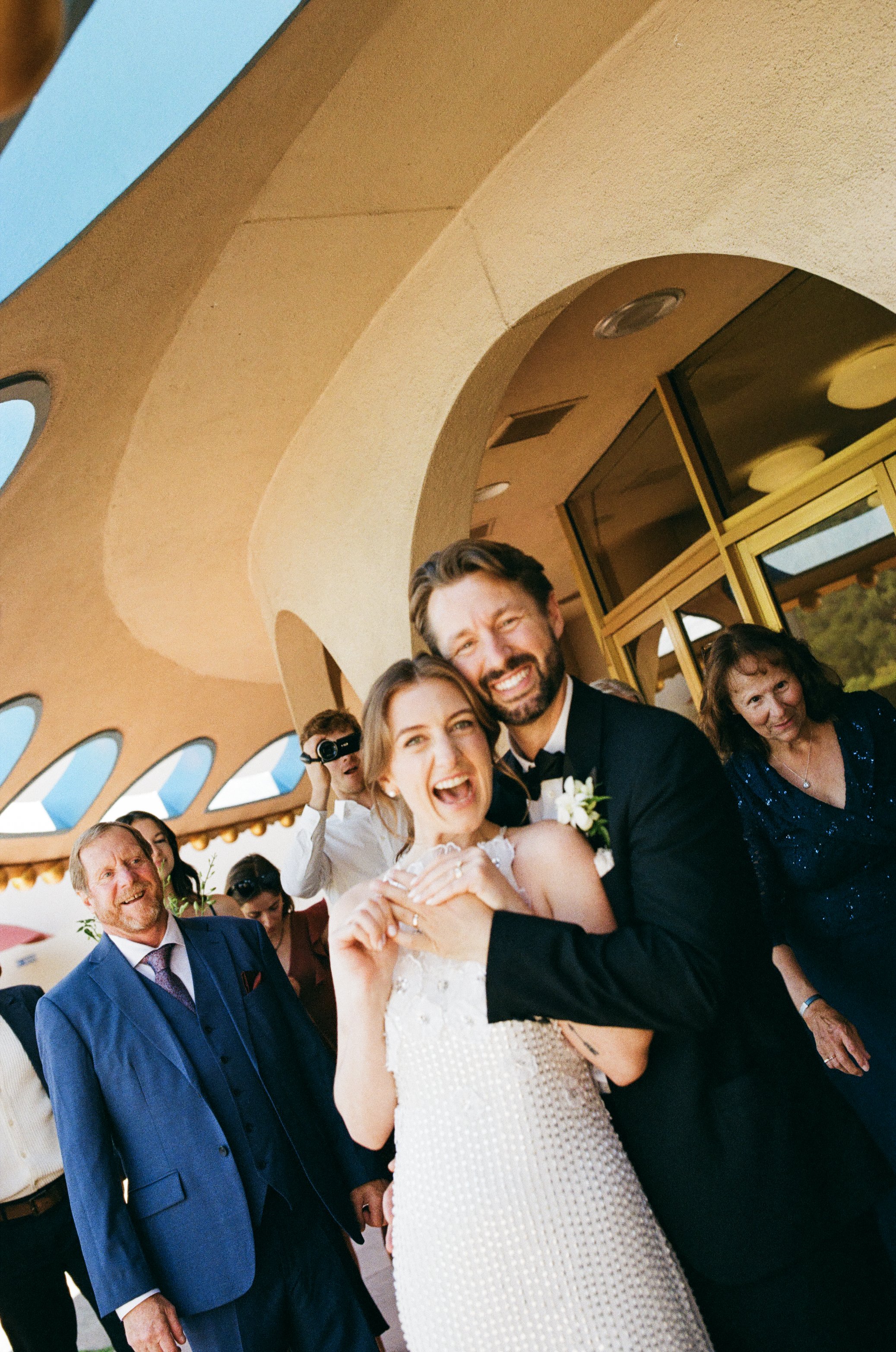 Smiling man in a blue suit at a wedding celebration with group of people, including a woman in a white dress and a man in a black tuxedo, under a decorative arch.