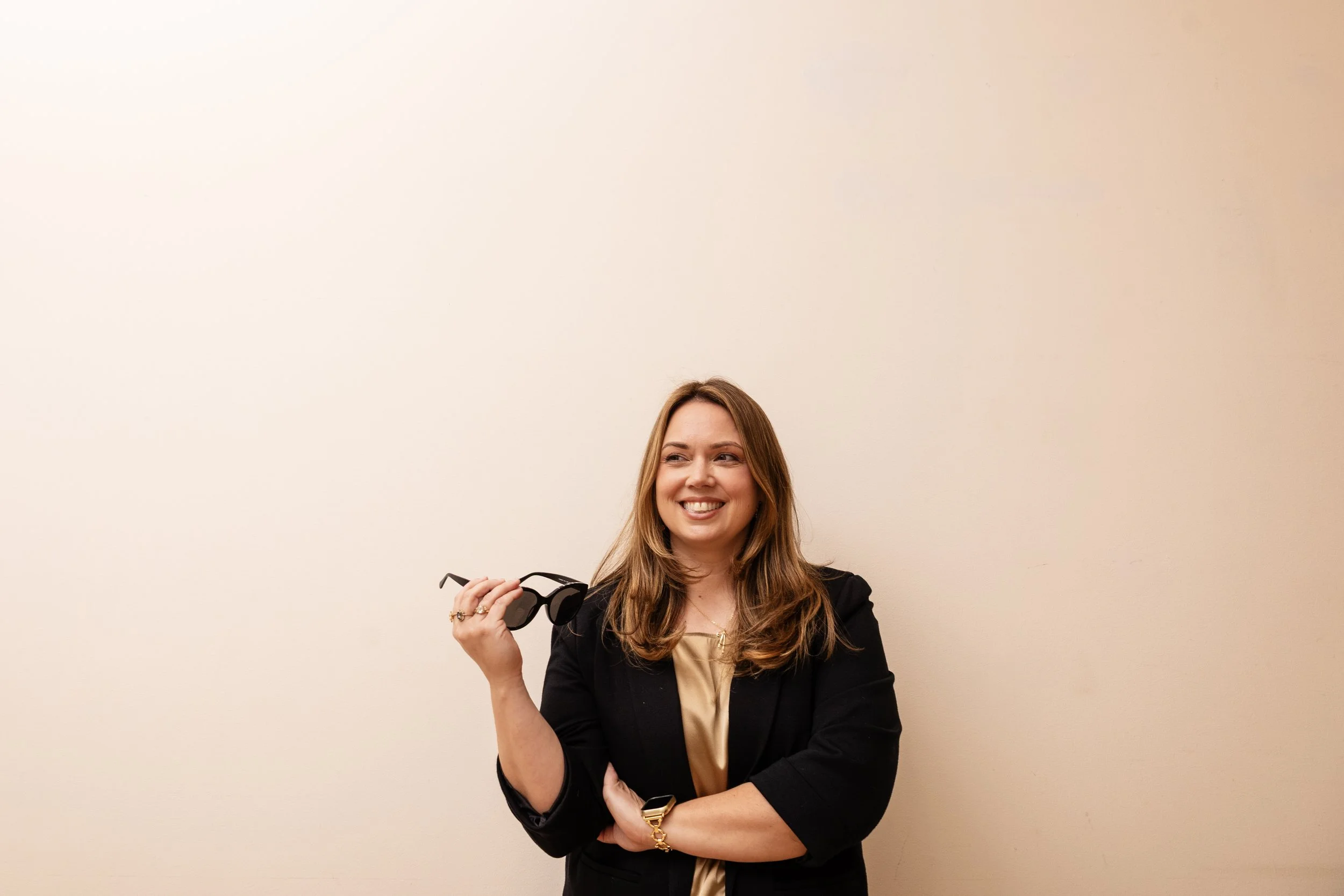 A woman with long brown hair holding sunglasses, smiling, standing against a plain beige wall.