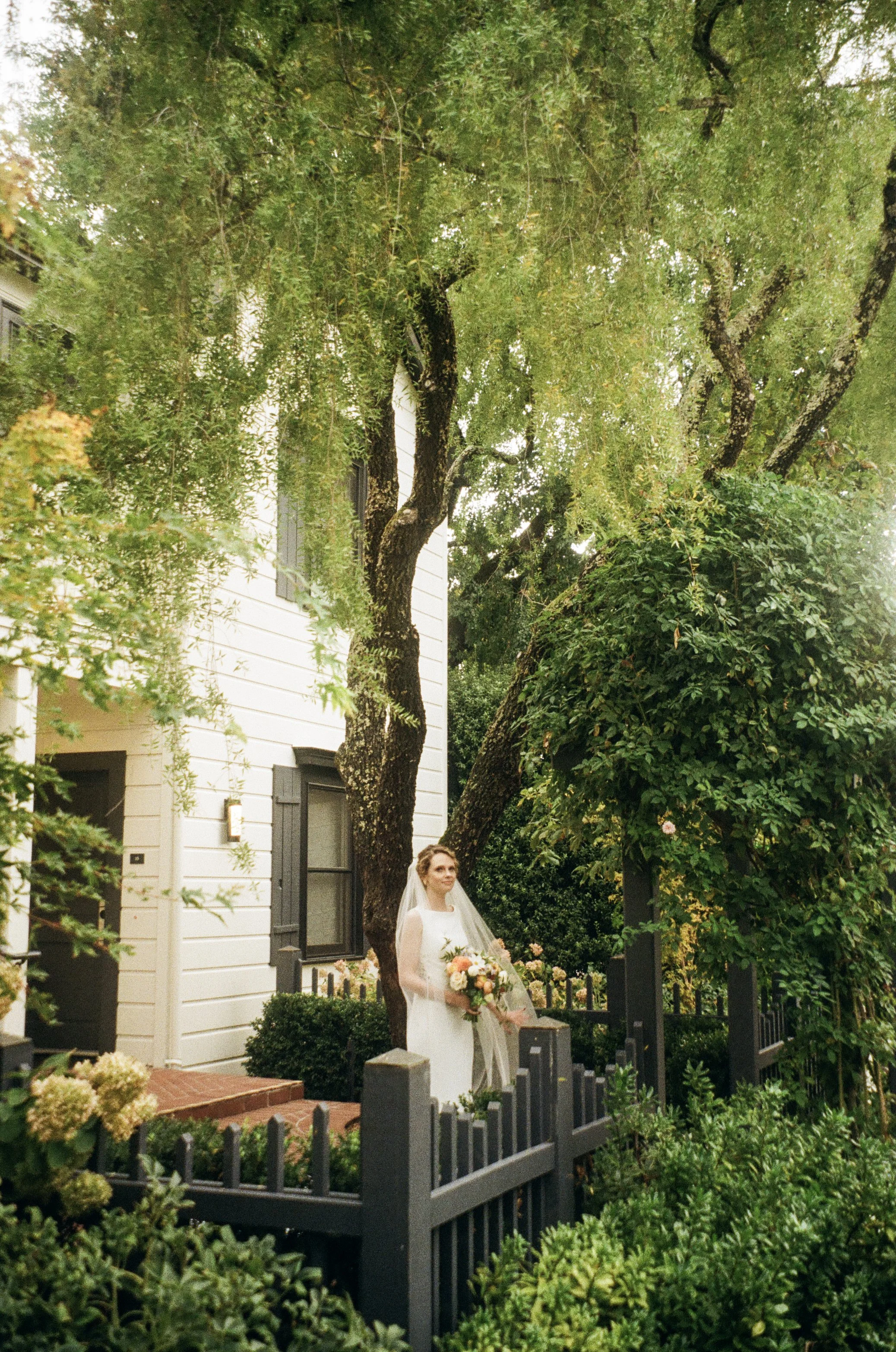 A bride in a white wedding dress holding a bouquet of flowers standing in a garden next to a black fence, with trees and a white house in the background.
