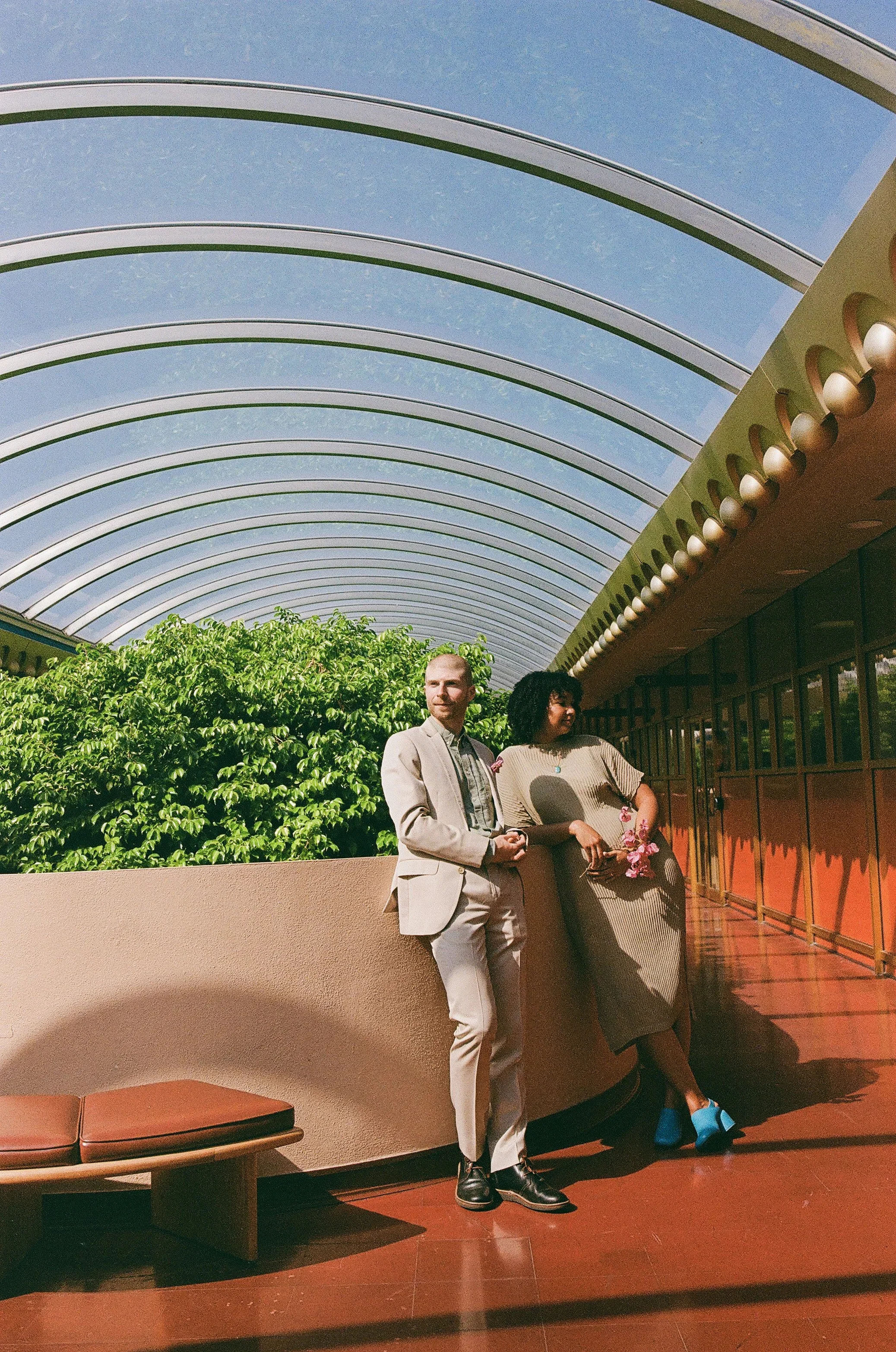 Two people, a man and a woman, are standing next to each other indoors with a futuristic arched glass ceiling above. The man is dressed in a beige suit, and the woman is wearing a beige dress with blue shoes and holding pink flowers. They are standing near a curved beige wall with greenery behind them and a red-tiled floor.