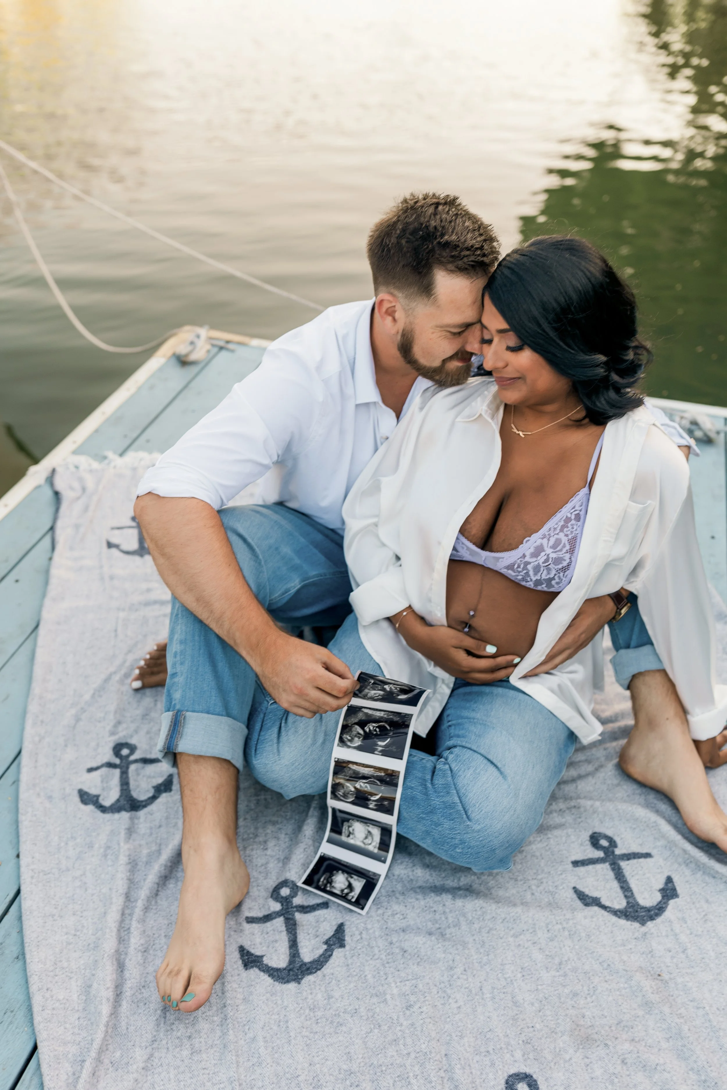 A couple sitting on a boat with mirrors, holding ultrasound images, touching foreheads and smiling, on a boat with an anchor design on the mat, near water.