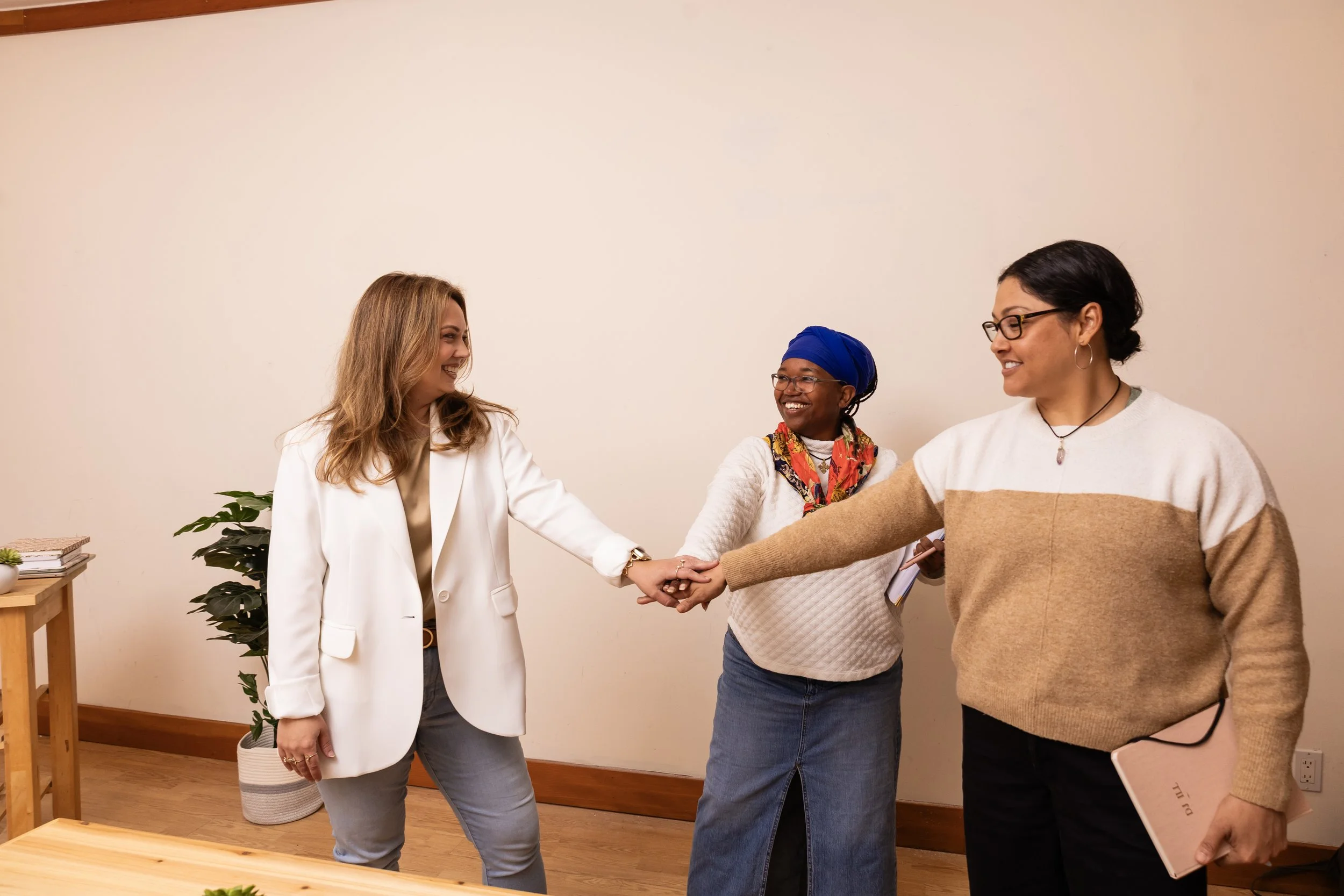 Three women standing in a room, holding hands, smiling, and facing each other. One woman has blonde hair, a white blazer, and grey pants. The second woman has dark hair, glasses, a colorful scarf, and a white sweater. The third woman has black hair, 