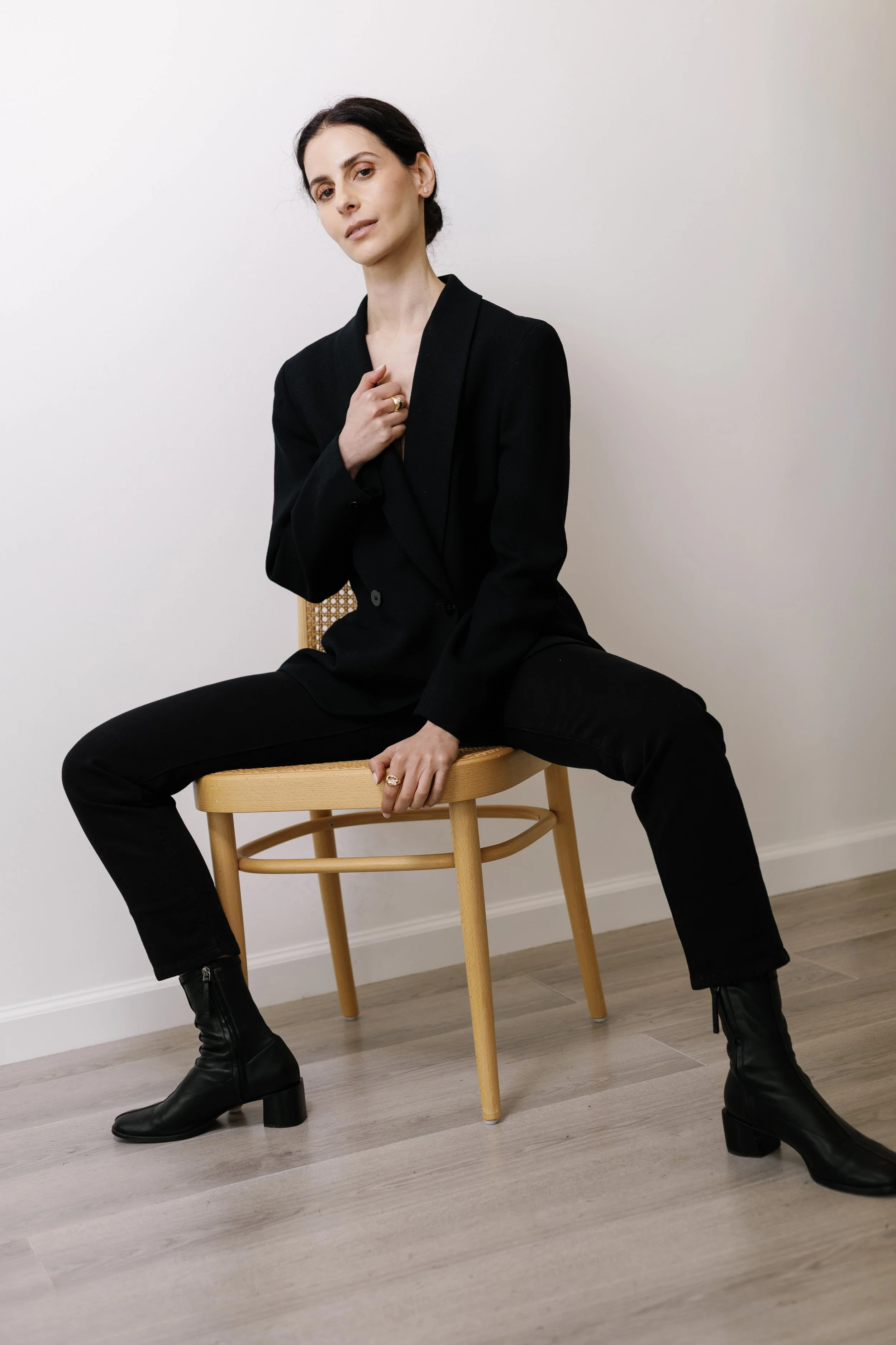 A woman with dark hair styled in an updo, wearing a black blazer, black pants, and black heeled boots, sitting on a wooden chair against a plain white wall, holding her blazer collar with one hand and resting her other hand on her knee.